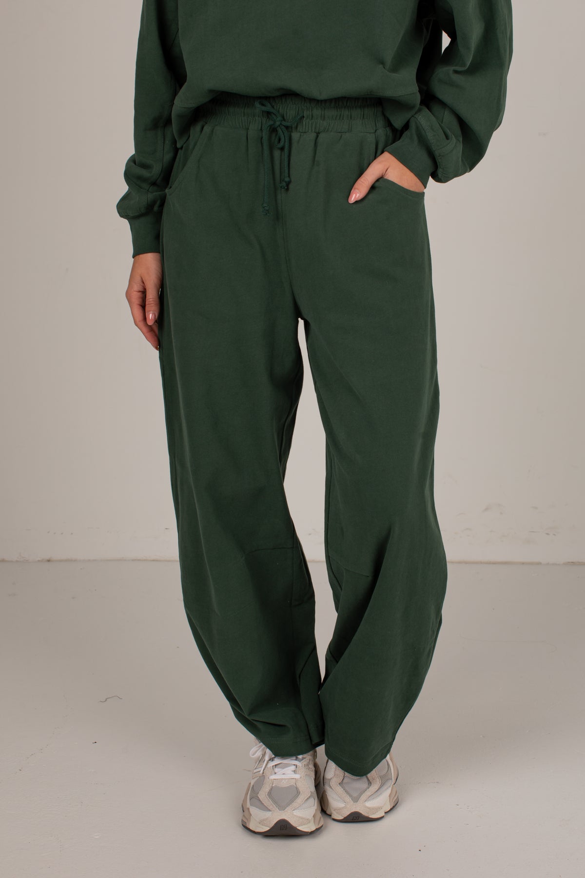 A person wearing dark green drawstring sweatpants with a matching sweatshirt and white sneakers, standing against a plain light gray studio background.