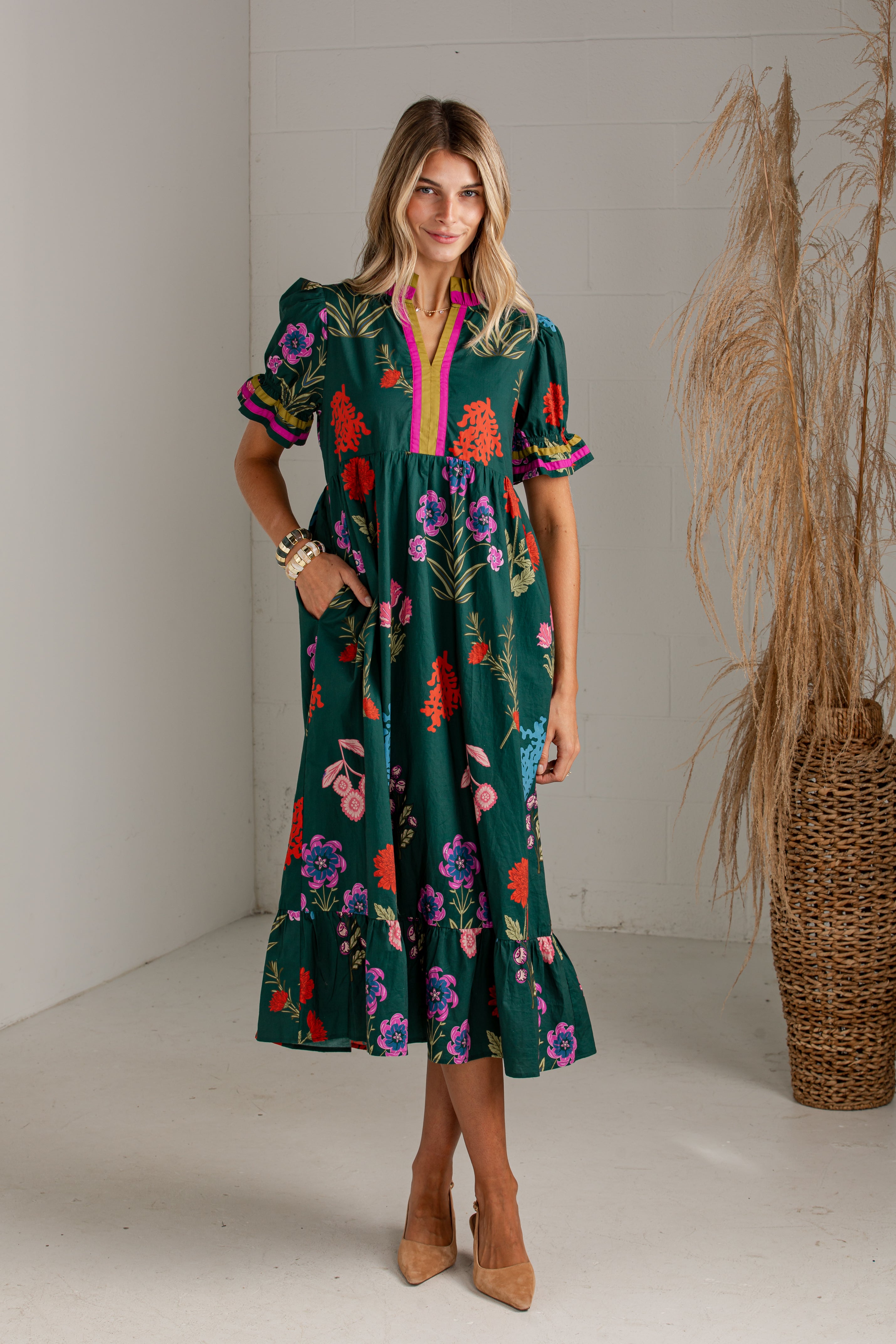 A woman models a dark green midi dress with a colorful floral print, short puff sleeves, and a bright pink collar, standing in a studio with a pampas grass vase in the background.
