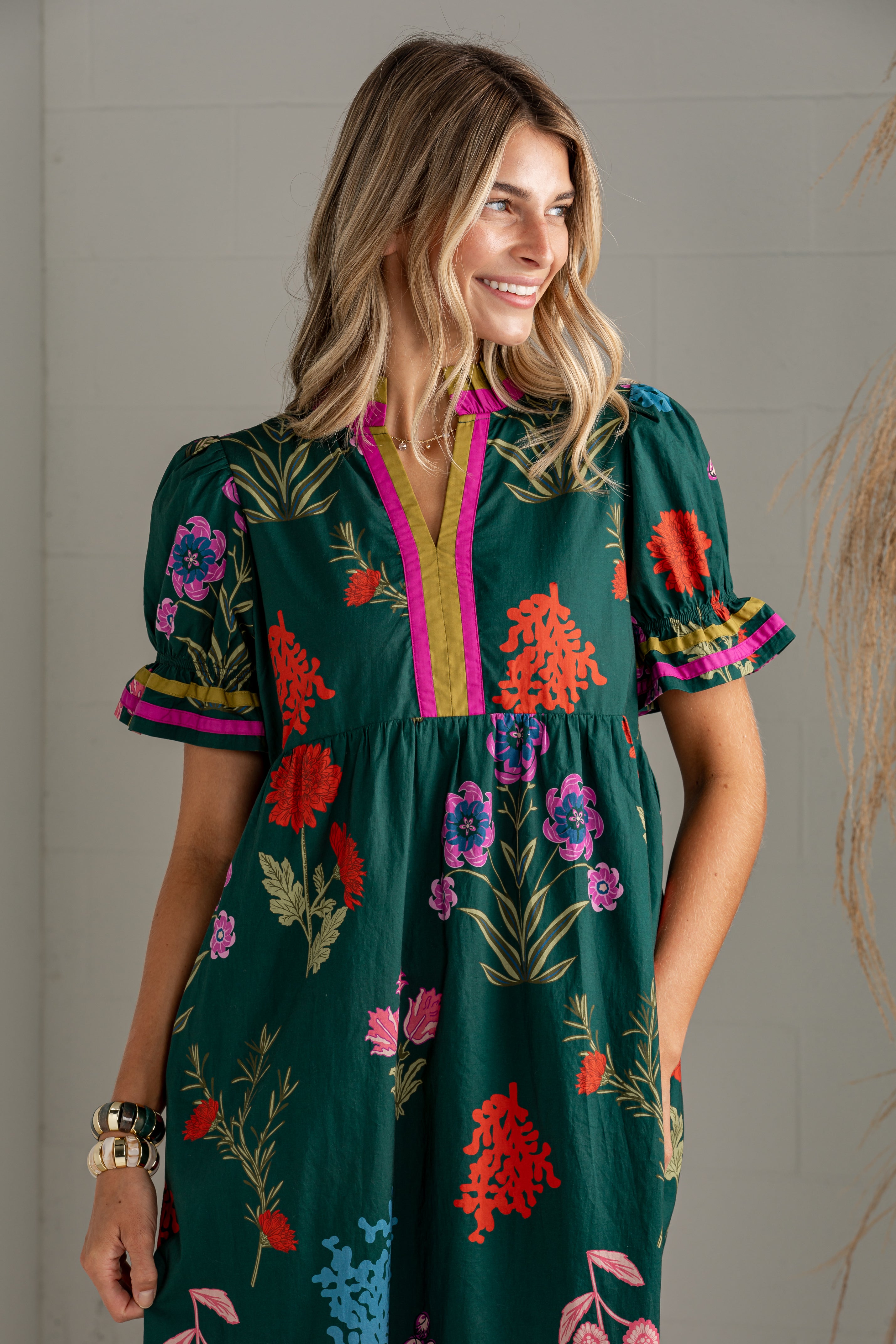 A woman wearing a dark green short-sleeve dress with a colorful floral print, pink trim, and puff sleeves, standing against a light gray wall.