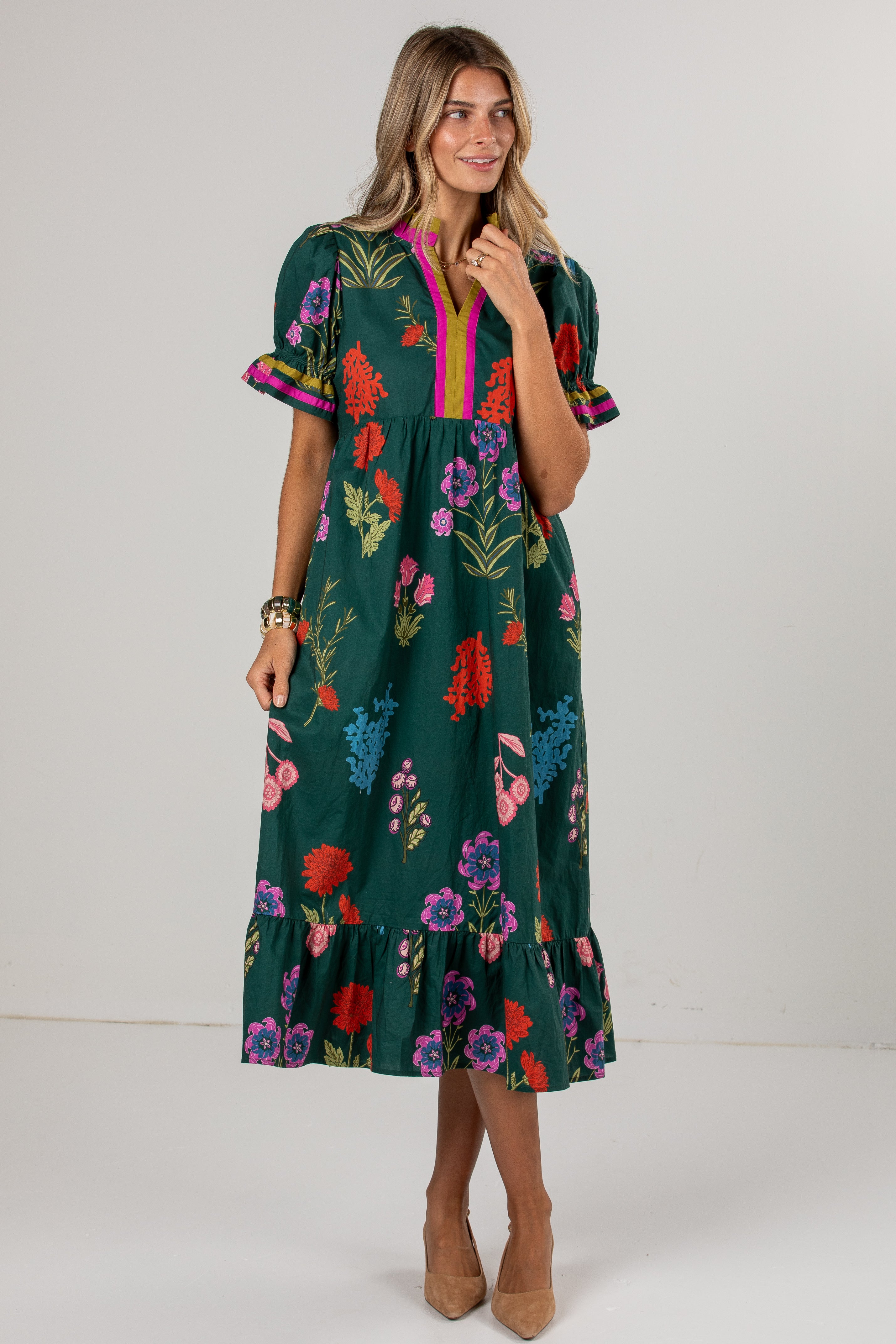 A woman modeling a dark green midi dress with a colorful floral print, puff sleeves, and a pink and green V-neckline, standing against a plain light gray studio background.