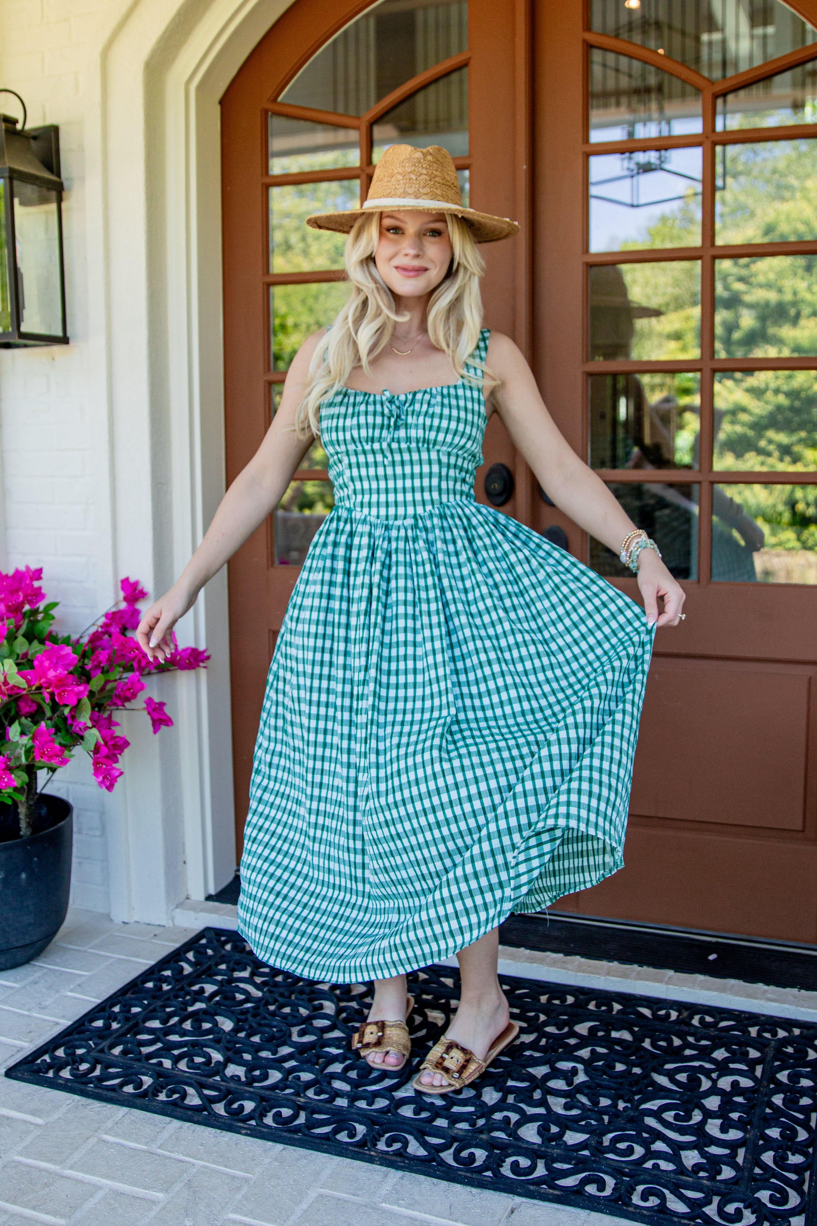 A woman wearing a green and white gingham midi dress with a straw hat stands on a front porch holding out her dress to show the flow. The porch has a brown wooden door, a black patterned doormat, and a potted plant with bright pink flowers.