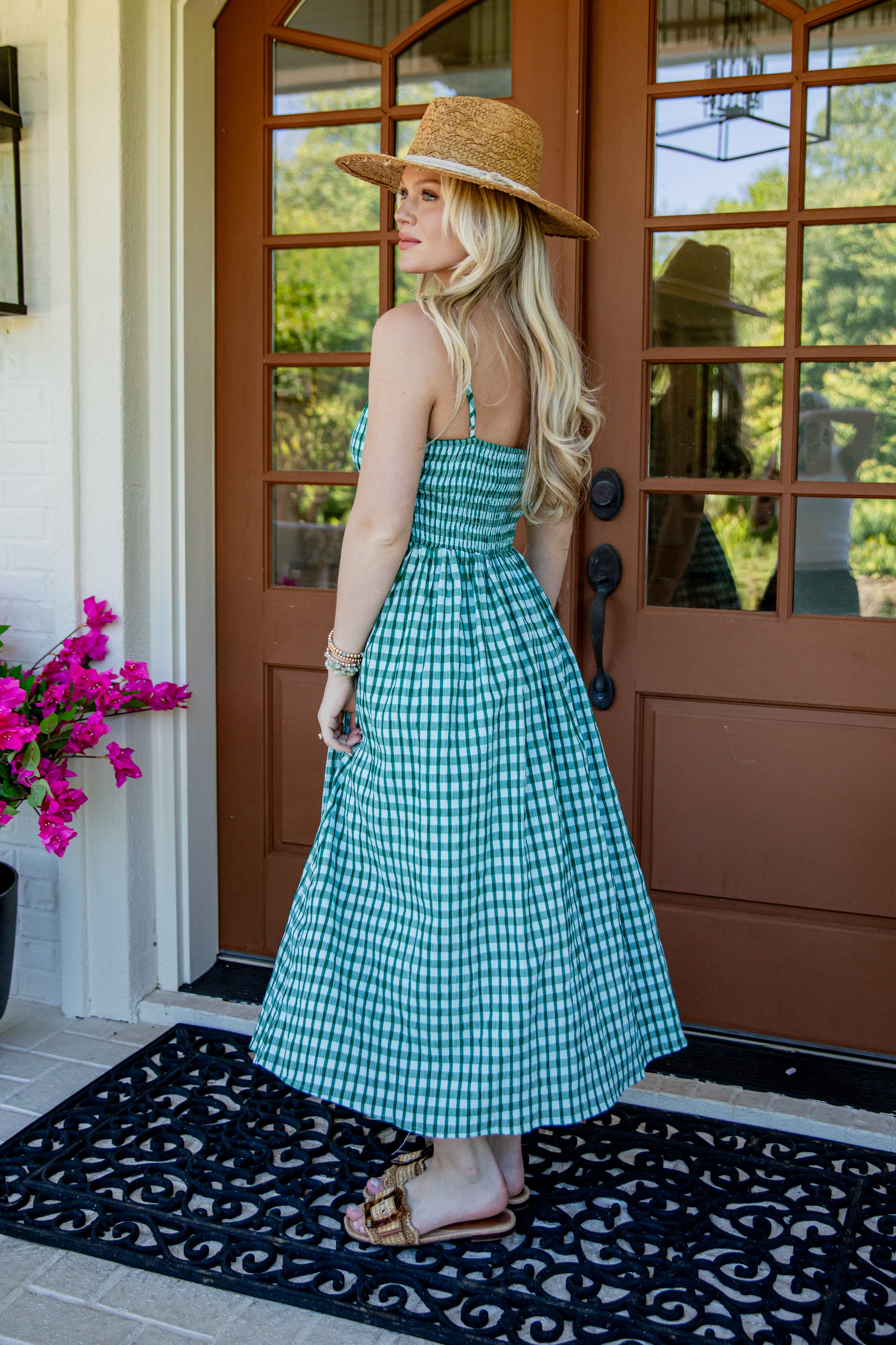 A woman stands on a front porch wearing a green and white gingham midi dress with a straw hat and sandals, photographed from the back.