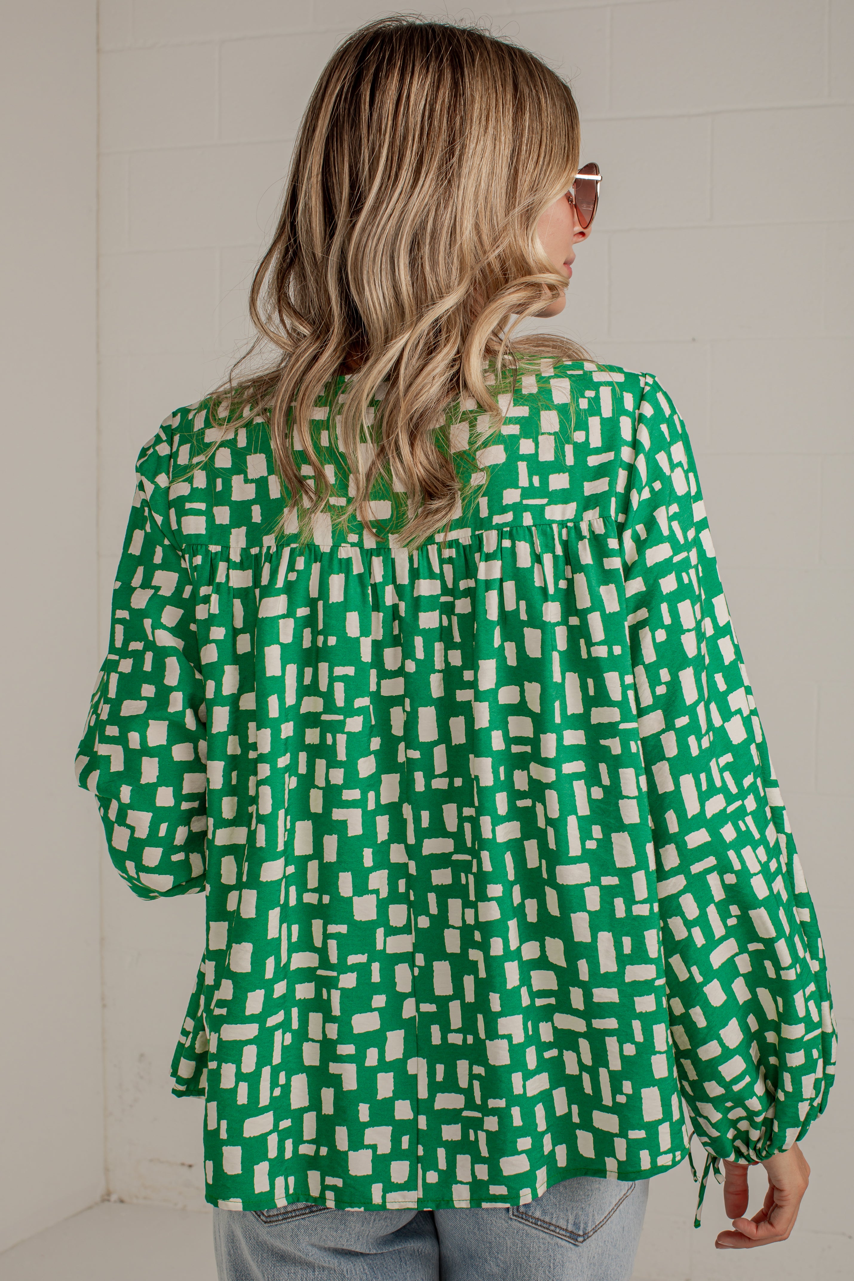 A woman viewed from behind wearing a green and white geometric patterned blouse with long sleeves and drawstring cuffs, paired with light blue jeans against a white brick wall.