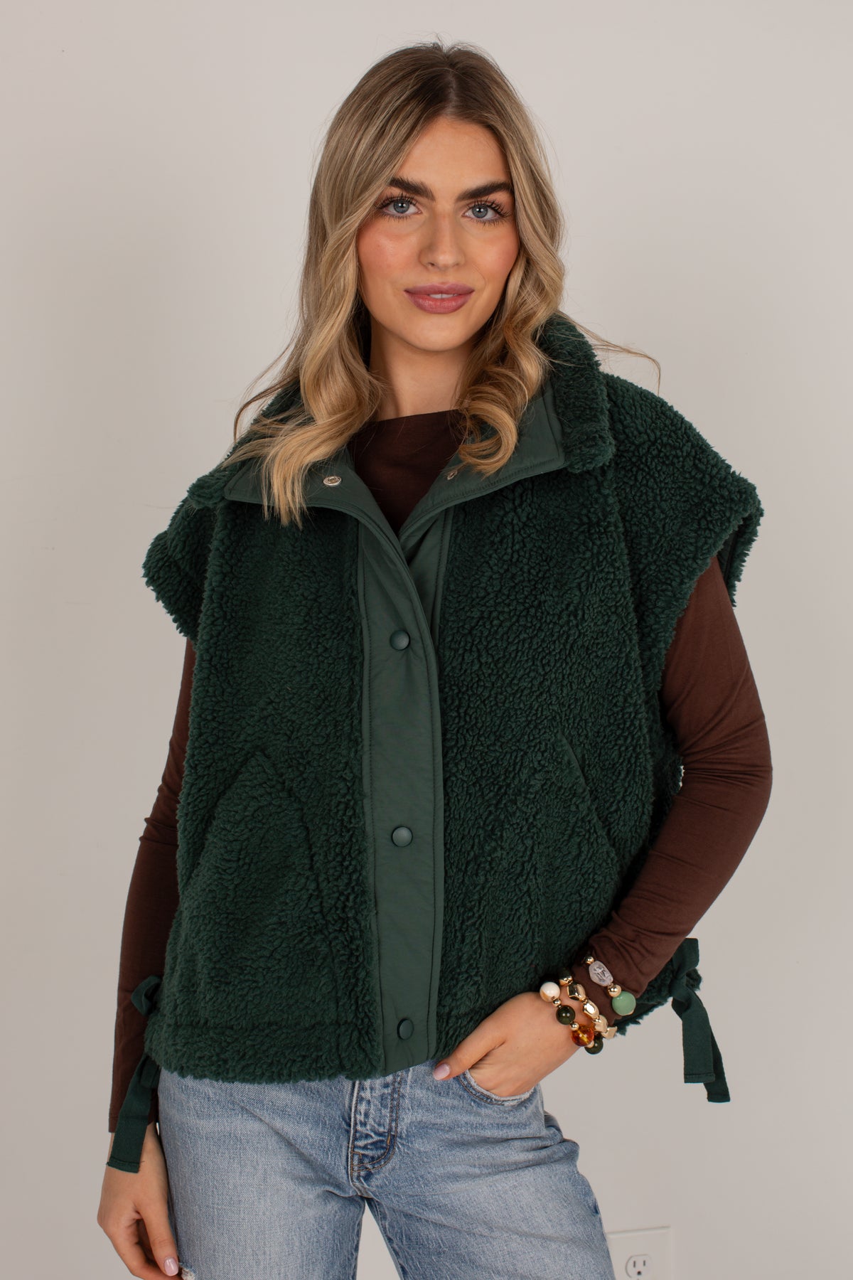 A woman modeling a dark forest green sherpa fleece vest with a button-front closure over a brown long-sleeve top and light-wash blue jeans, set against a plain light gray studio background.