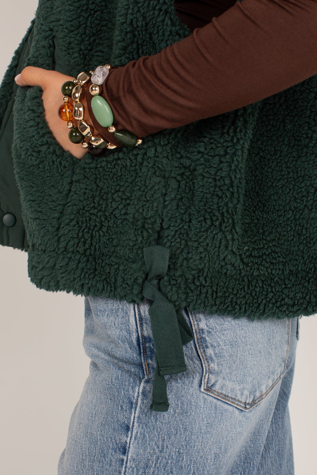 Close-up of a person wearing a dark forest green sherpa fleece vest over a brown long-sleeve top and light blue jeans, accessorized with multiple beaded bracelets.