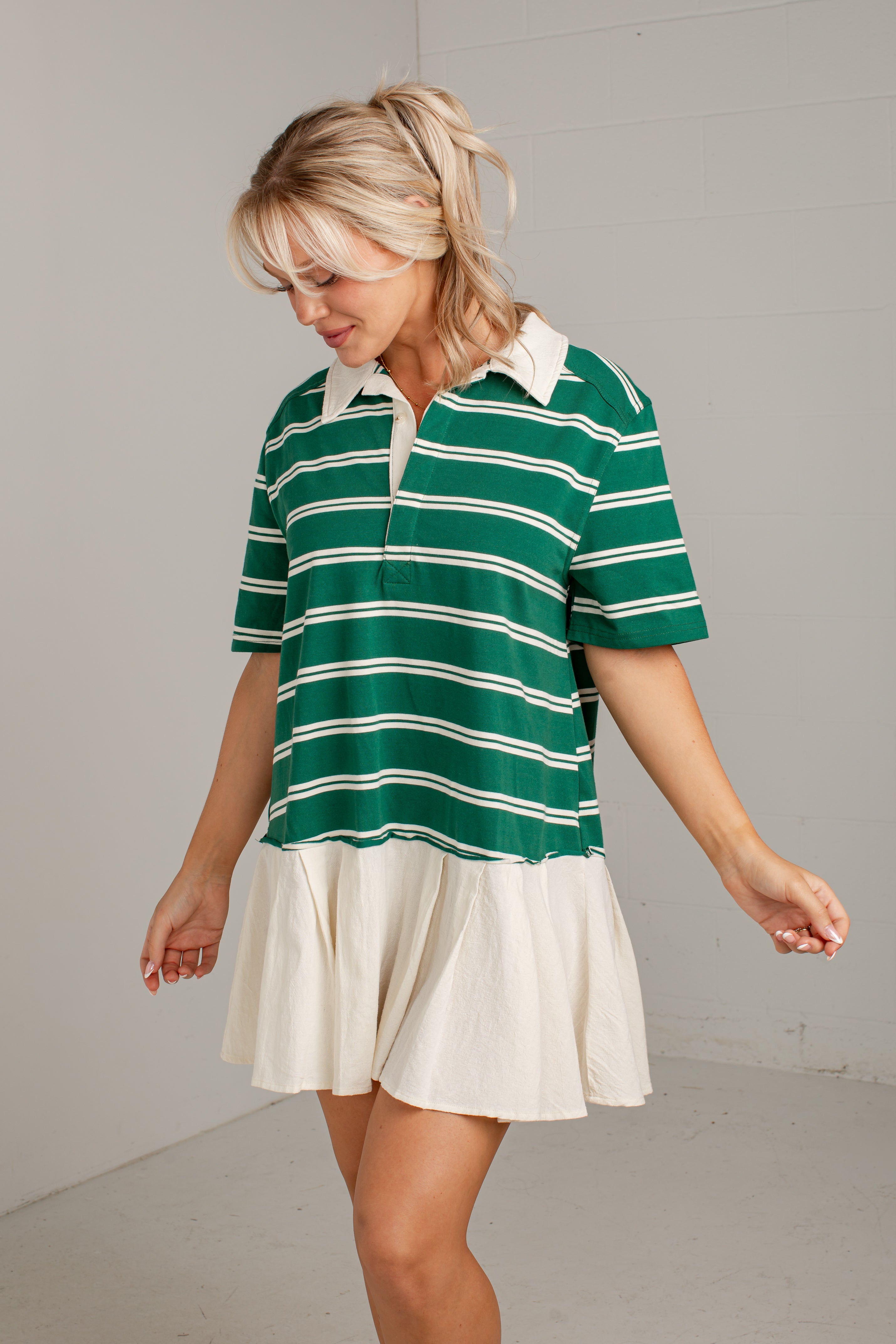 A woman wearing a green and white striped short-sleeve polo shirt with an off-white pleated mini skirt, standing against a light gray studio background.