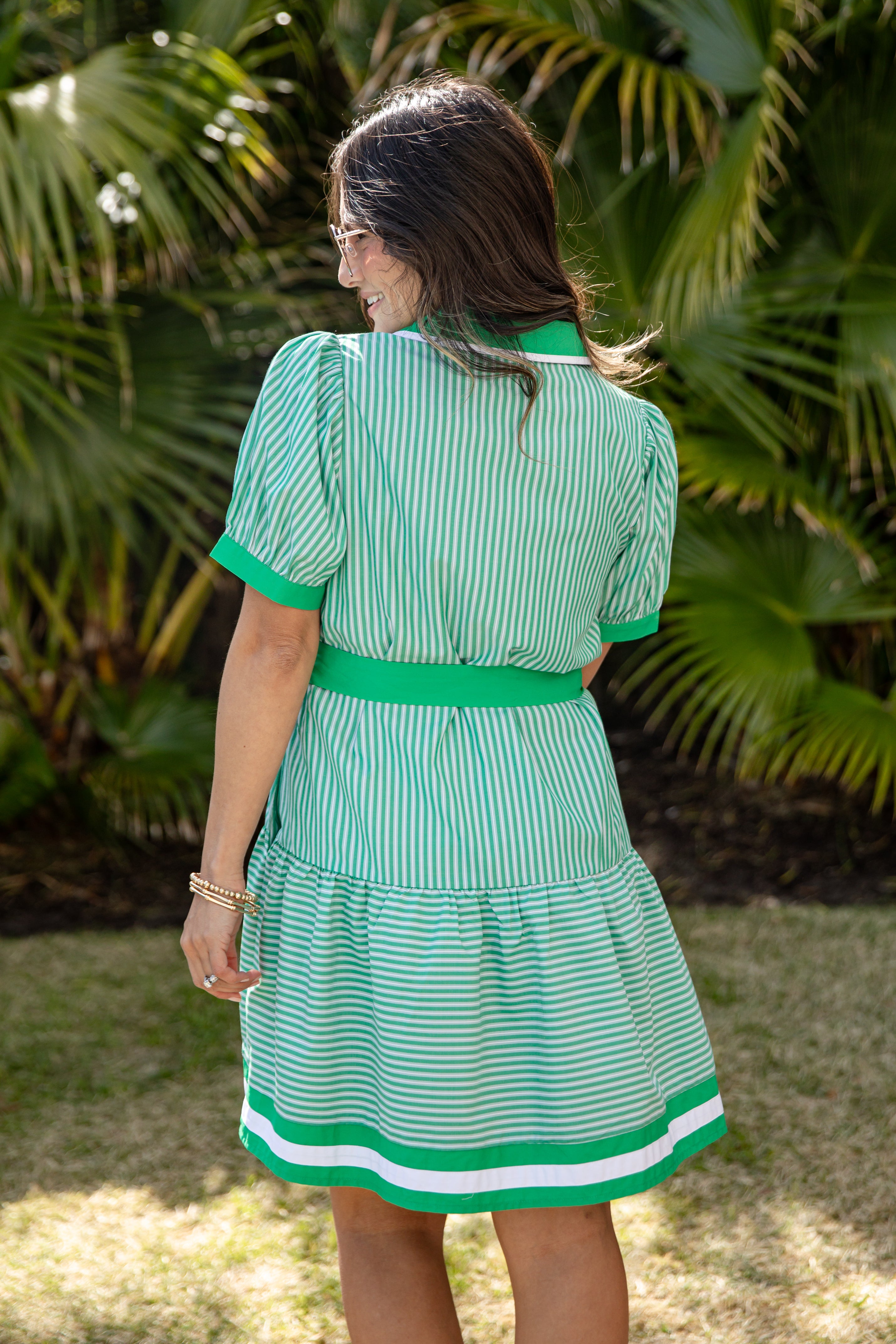 Woman wearing a green and white striped dress with a belt, standing in front of tropical plants.