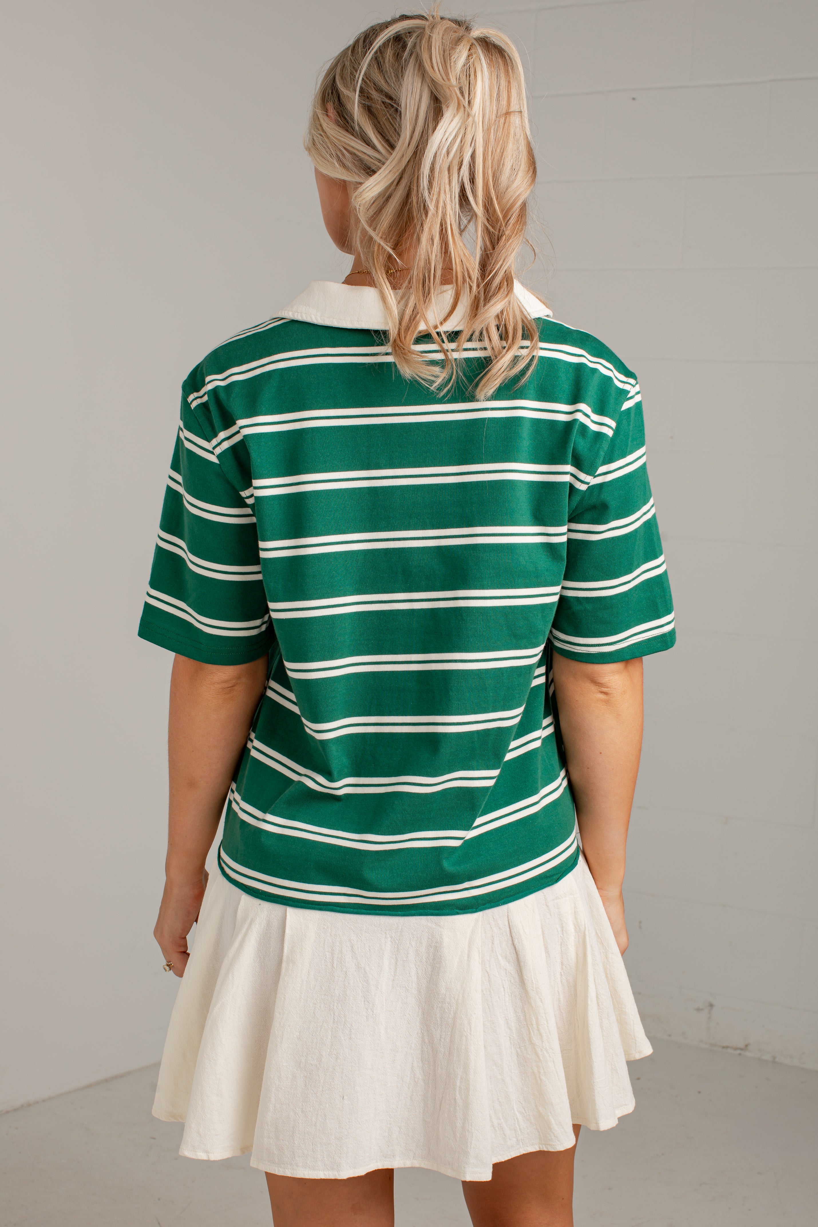 A woman viewed from behind wearing a green short-sleeve polo shirt with thin white horizontal stripes and a white pleated skirt, standing against a light gray wall.