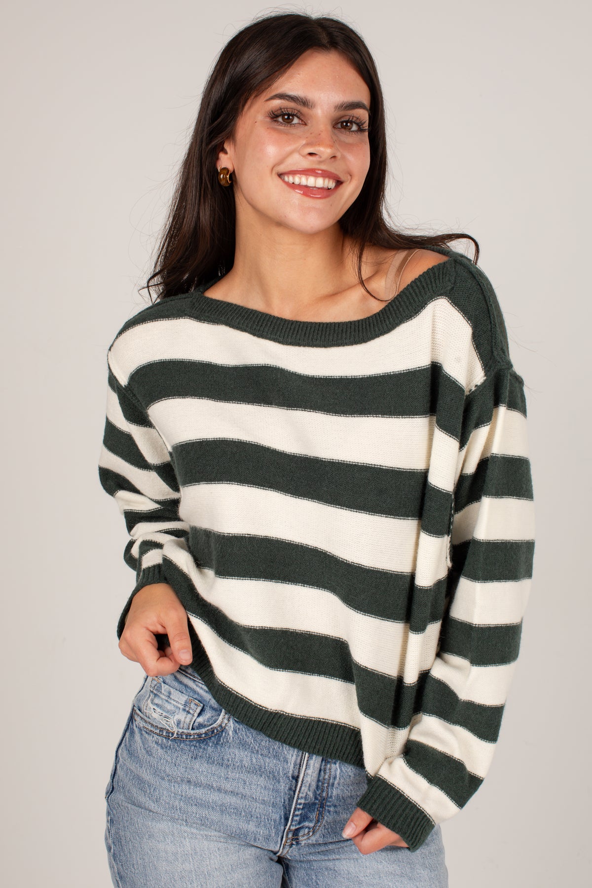 A woman wearing a dark green and cream striped knit sweater with an off-the-shoulder design, paired with light blue jeans, against a plain light gray background.