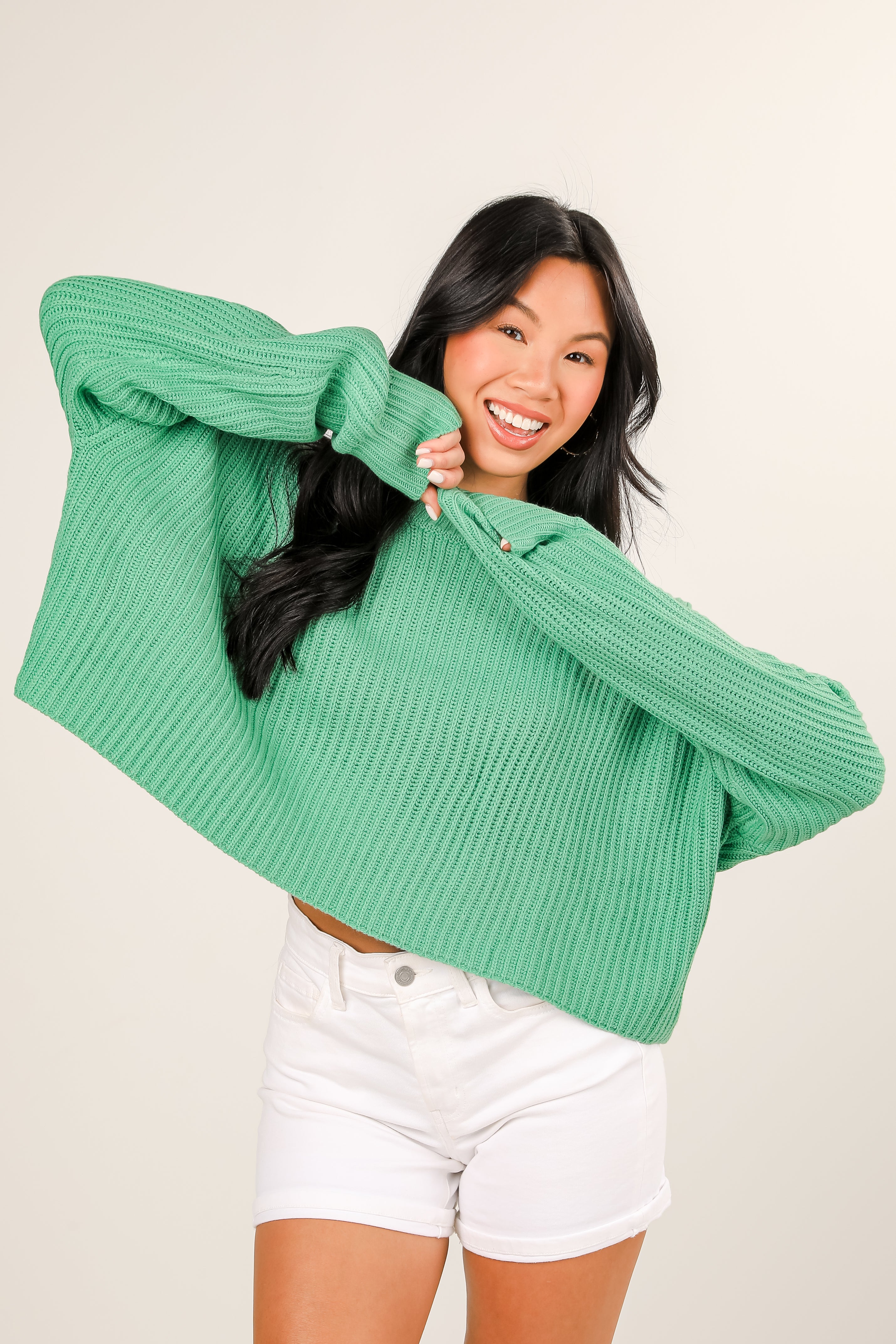 Woman wearing a green sweater and white shorts against a beige background