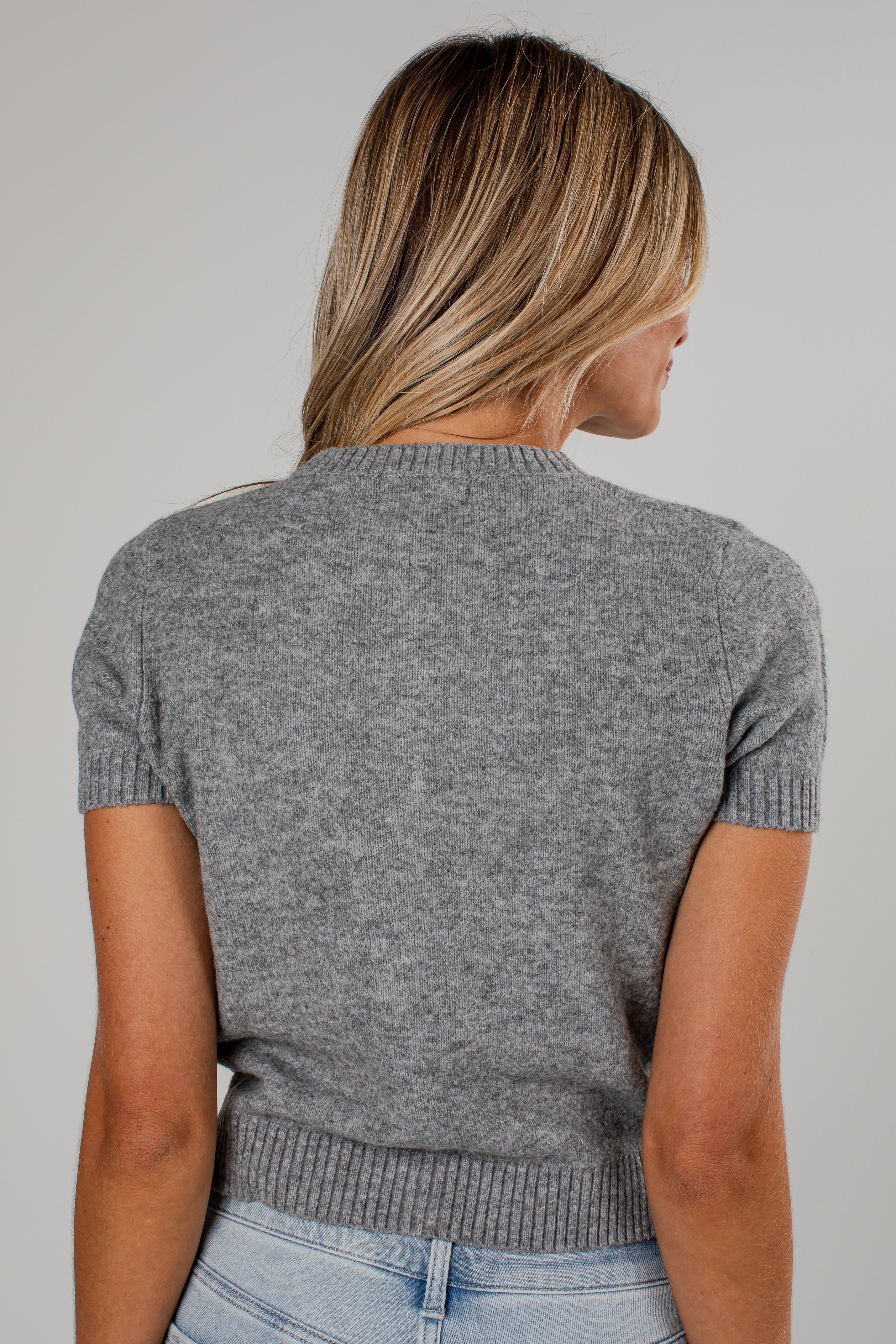 A woman viewed from behind wearing a short-sleeve gray knit sweater and light blue jeans against a plain light gray studio background.