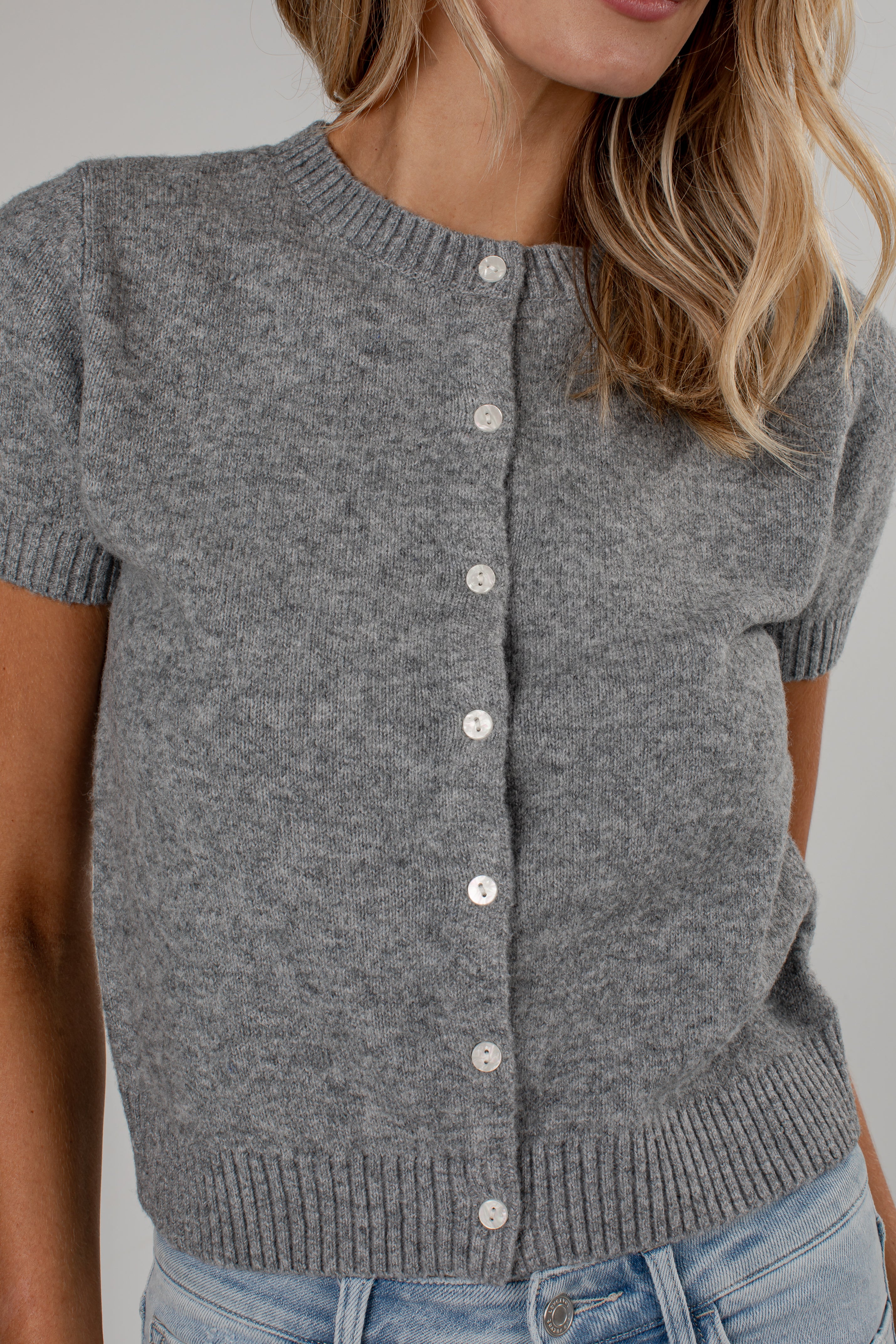 A close-up of a woman wearing a short-sleeve gray knit cardigan with white buttons, paired with light blue jeans, against a plain light gray background.