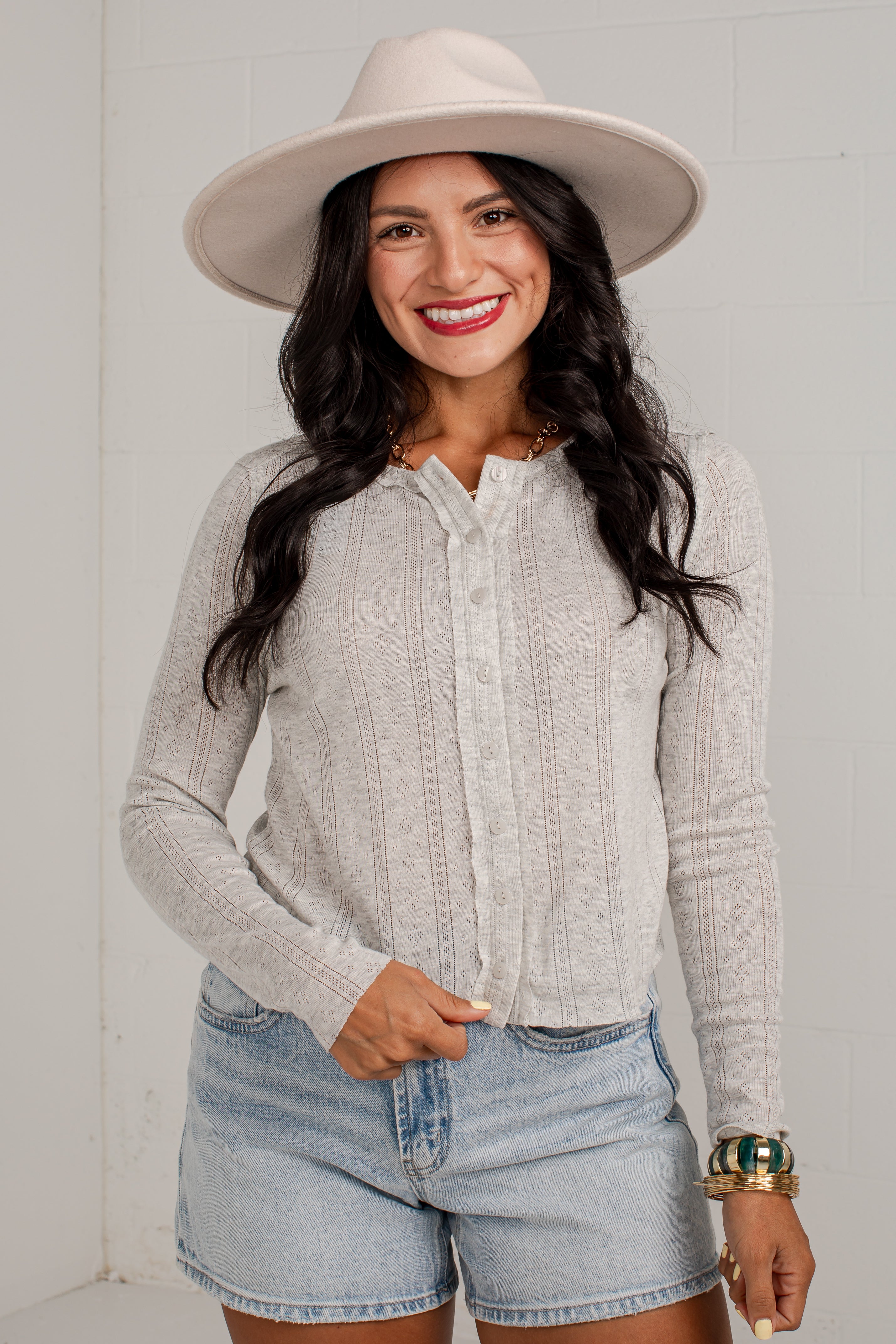A woman wearing a light beige wide-brim fedora hat, a light gray ribbed knit long-sleeve button-up shirt, and denim shorts, standing against a white brick wall.