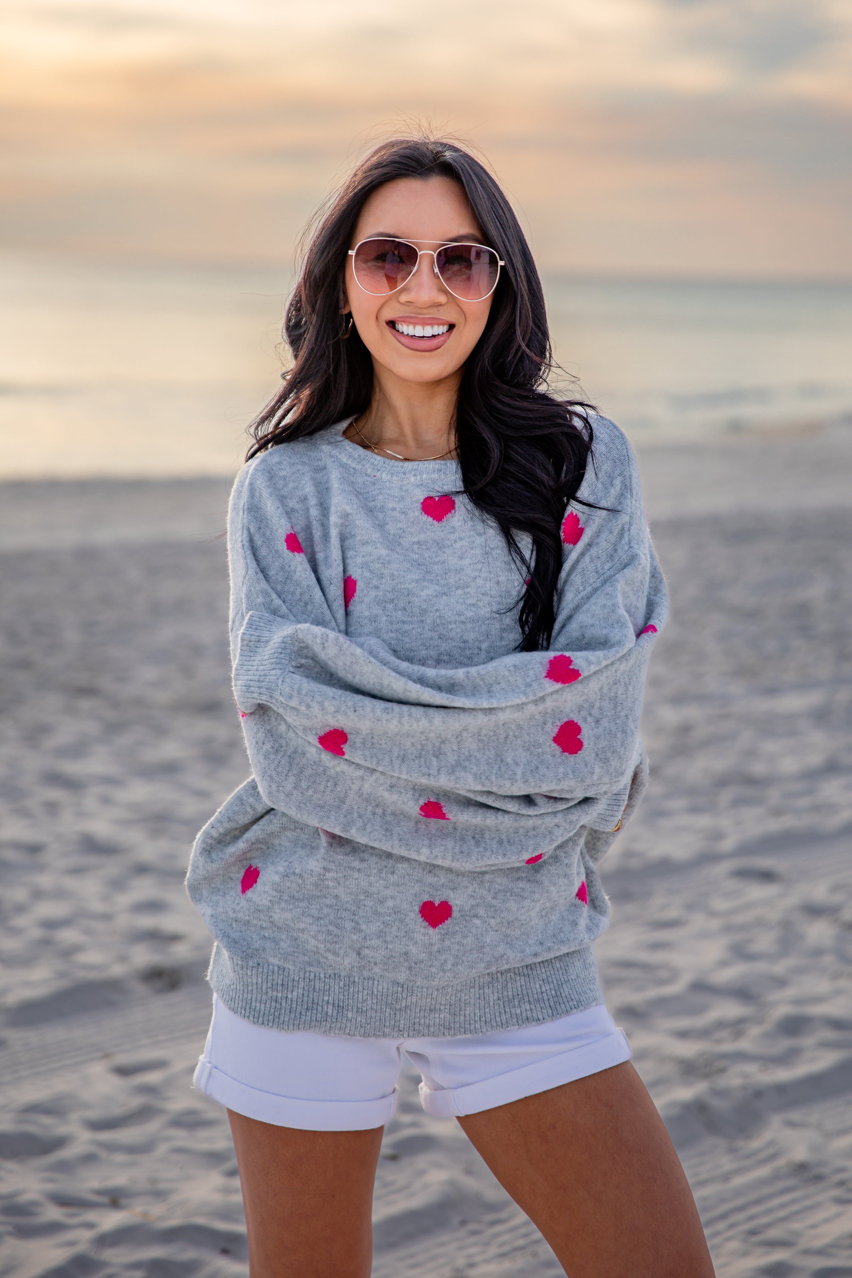 Woman wearing a gray sweater with red heart patterns on a beach at sunset.