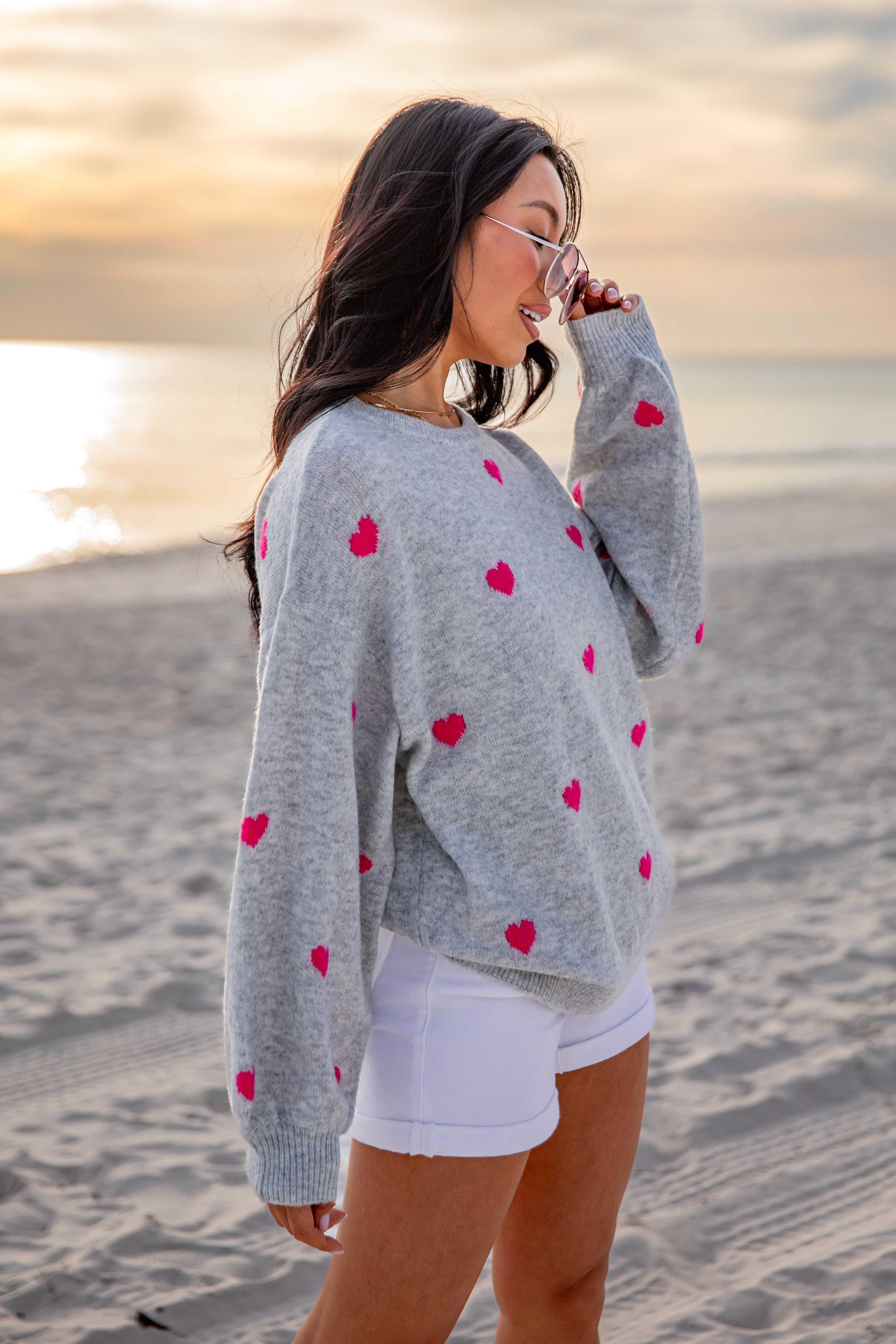 Woman wearing a gray sweater with red heart patterns on a beach at sunset.