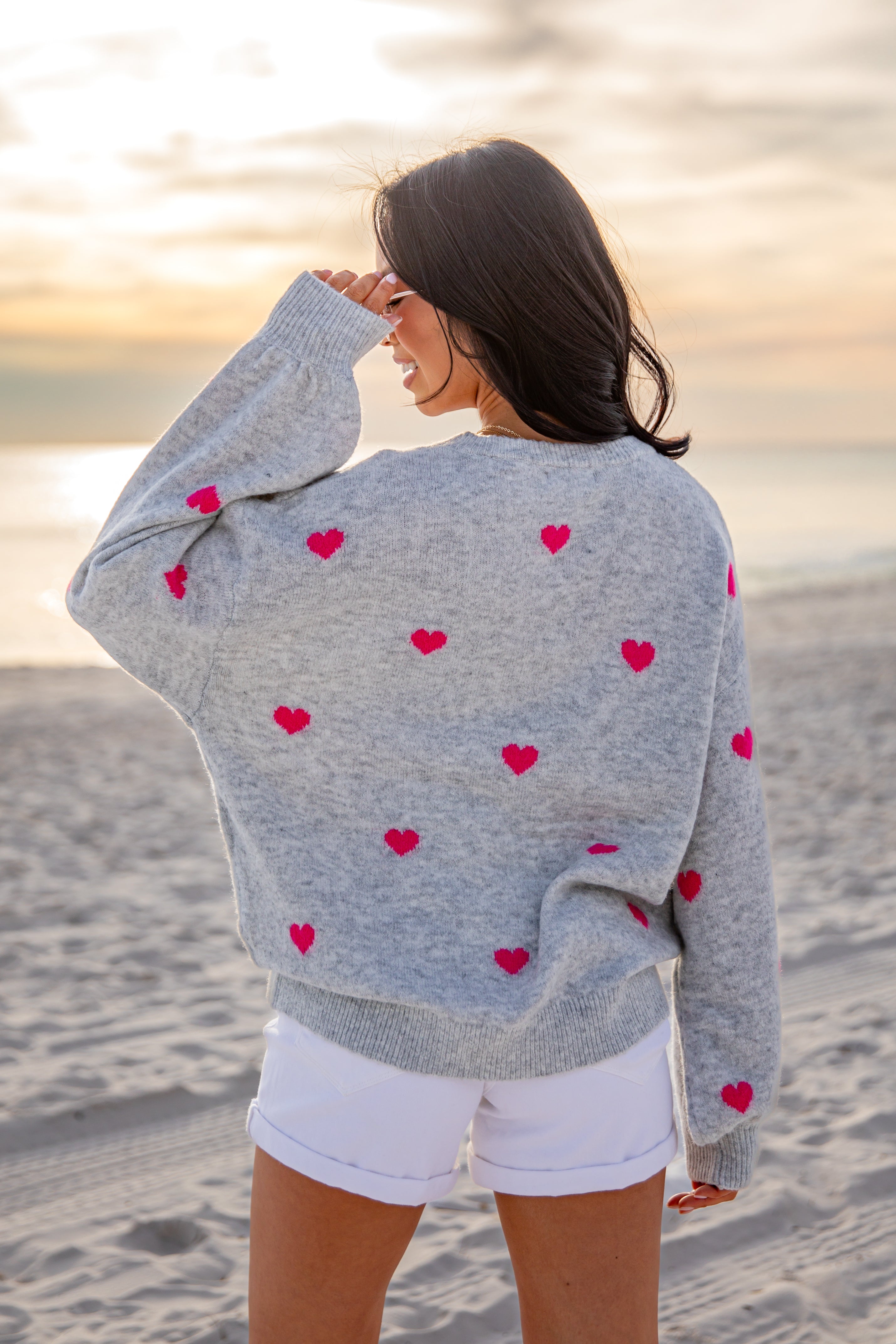 Woman wearing a gray cardigan with red heart patterns on a beach at sunset.