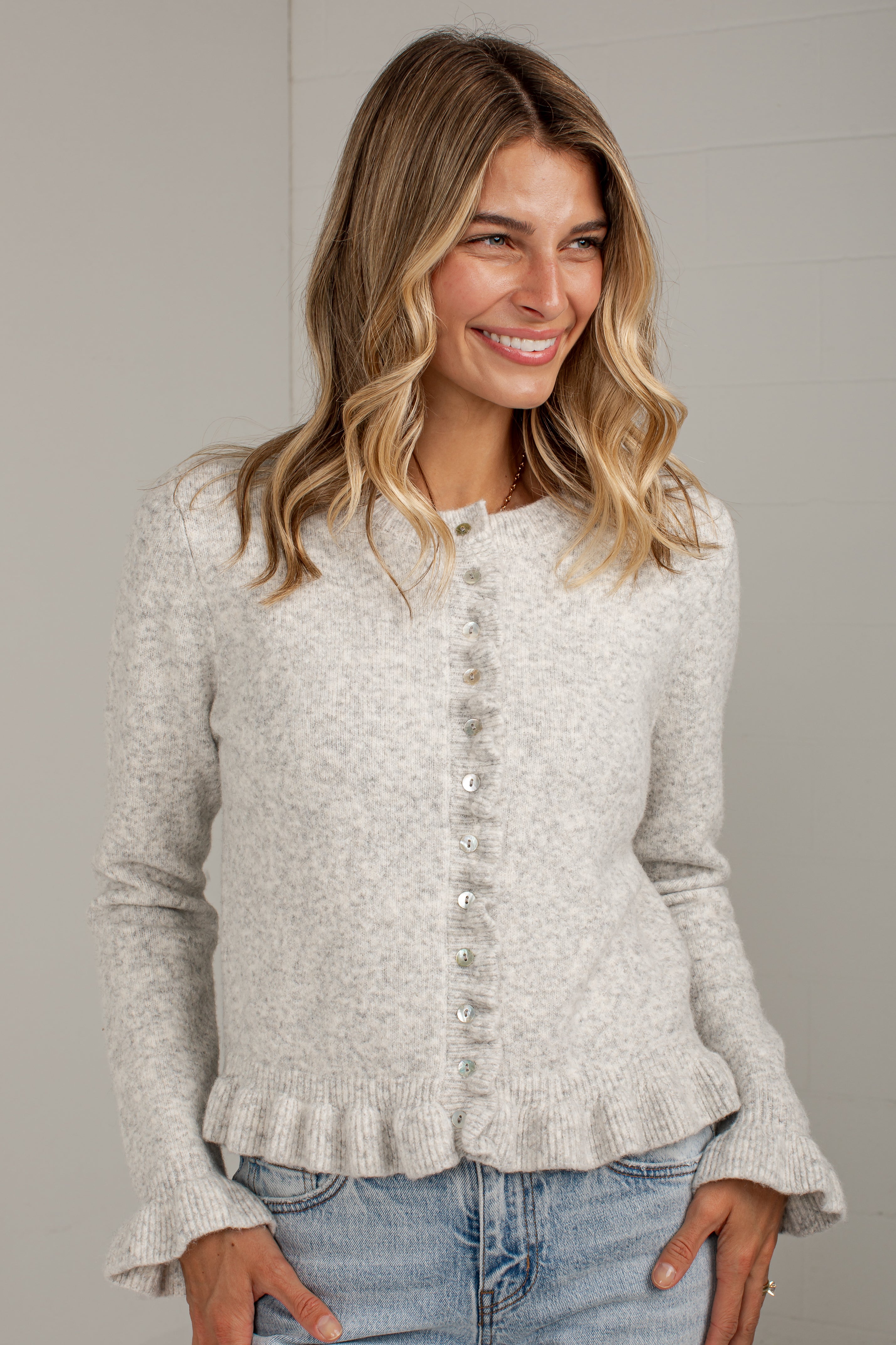 A woman wearing a light heather gray knit cardigan with a button-down front and ruffled hem, paired with blue jeans, against a neutral gray background.