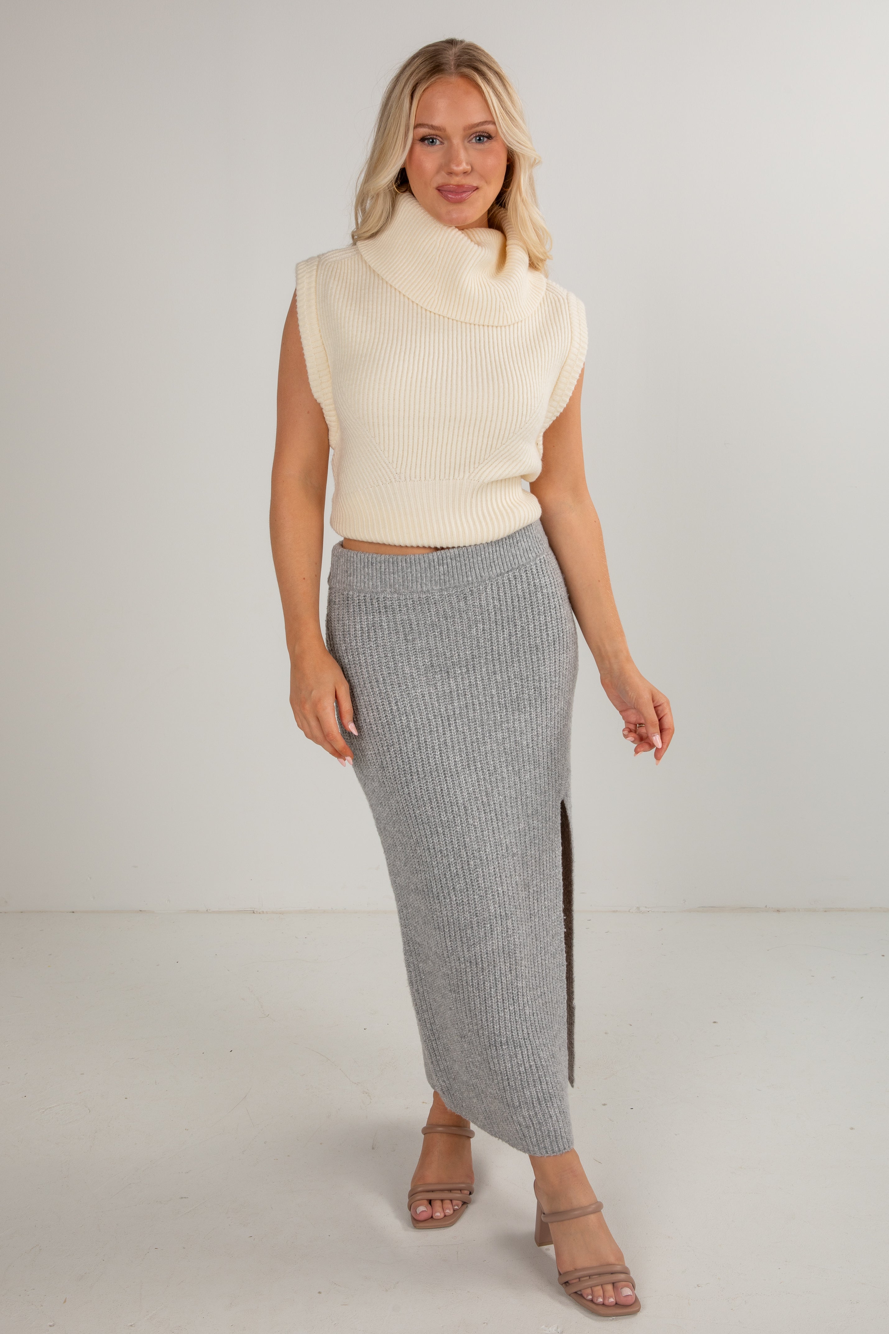 Woman wearing a cream sleeveless sweater and gray skirt on a white background