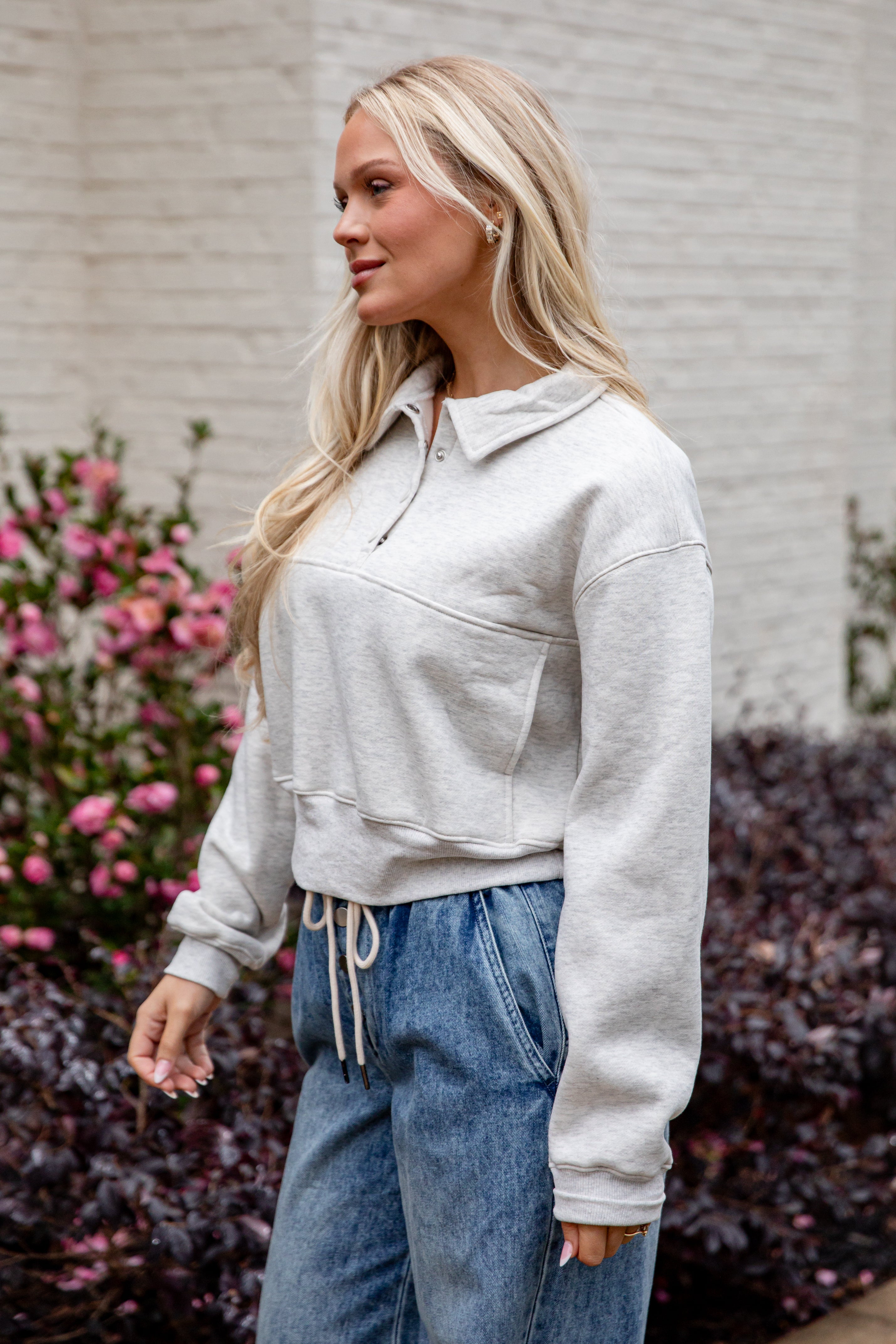 Woman wearing a light gray sweatshirt and blue jeans standing outdoors with flowers and a building in the background.