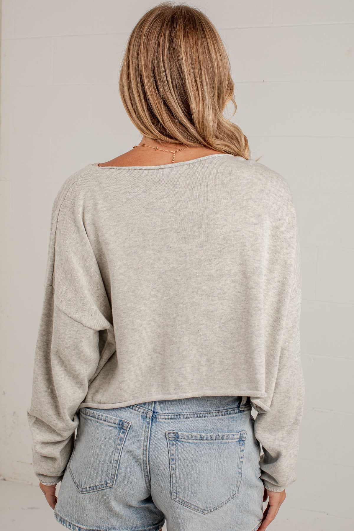 A woman viewed from behind wearing a light heather gray long-sleeve cropped sweater and light-wash blue jeans against a white paneled wall.