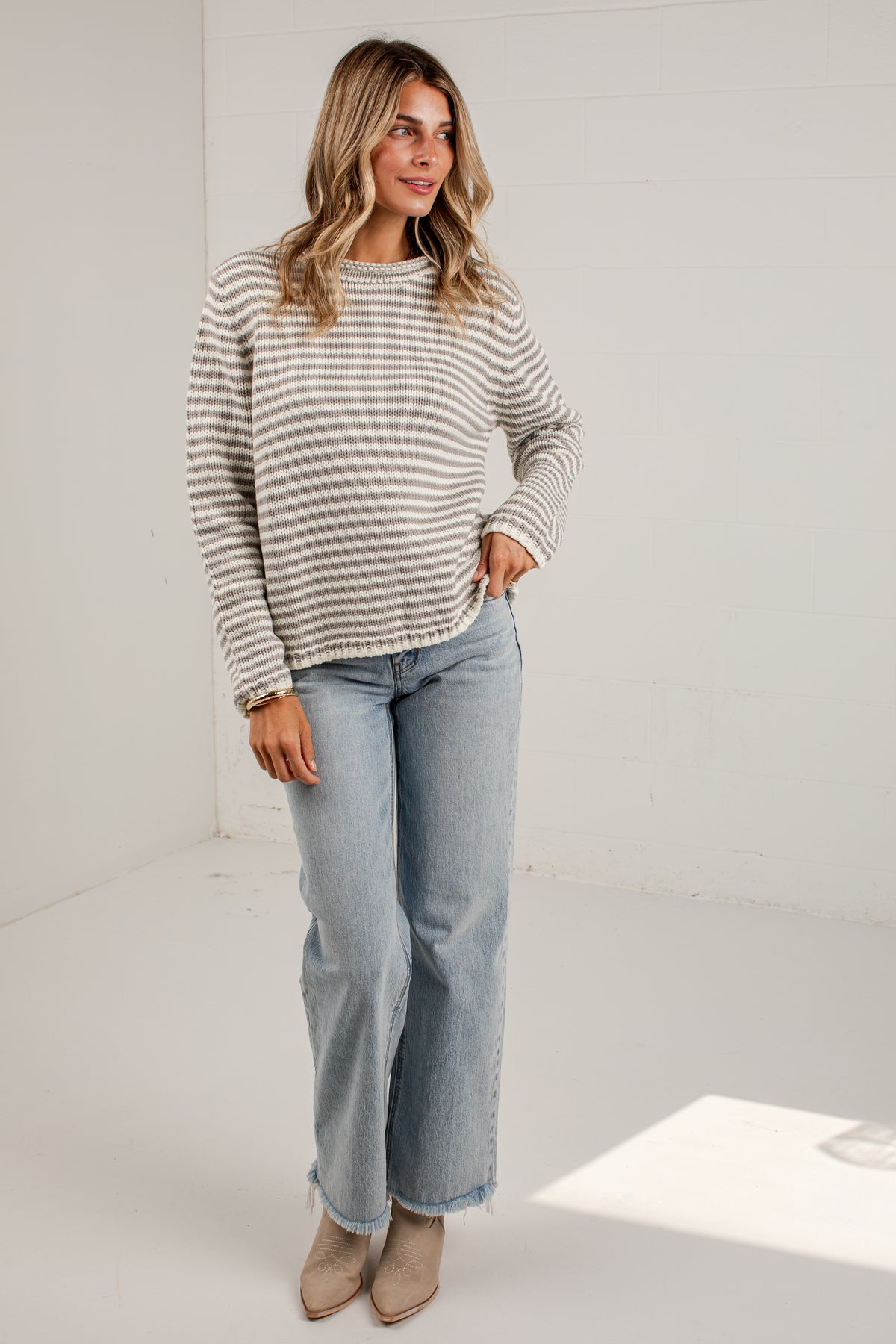 A woman modeling a beige and white horizontally striped knit sweater, paired with light-wash blue jeans and beige ankle boots, standing in a bright white studio setting.
