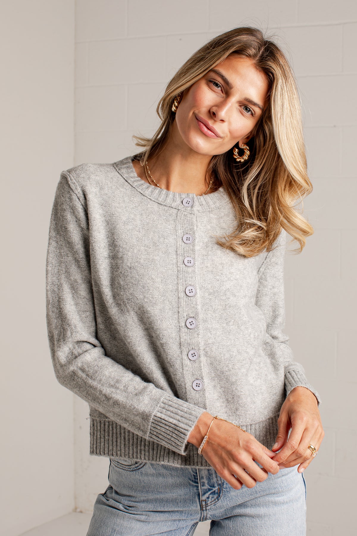 A woman wearing a light gray knit cardigan sweater with a button-down front, paired with light blue jeans, standing against a white tiled wall.