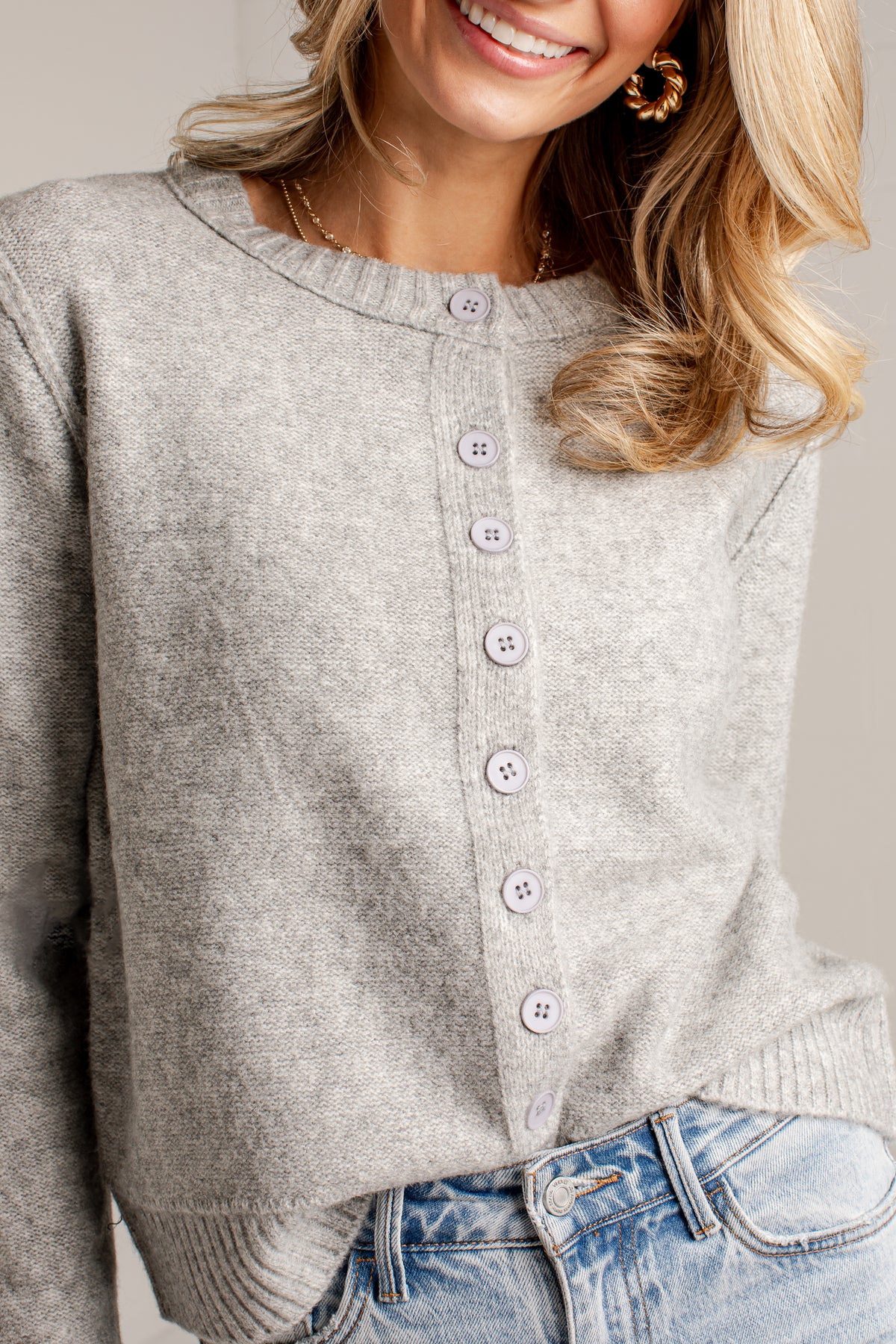 A close-up of a woman wearing a light gray heather knit cardigan with a button-front closure and light gray buttons, paired with blue jeans.
