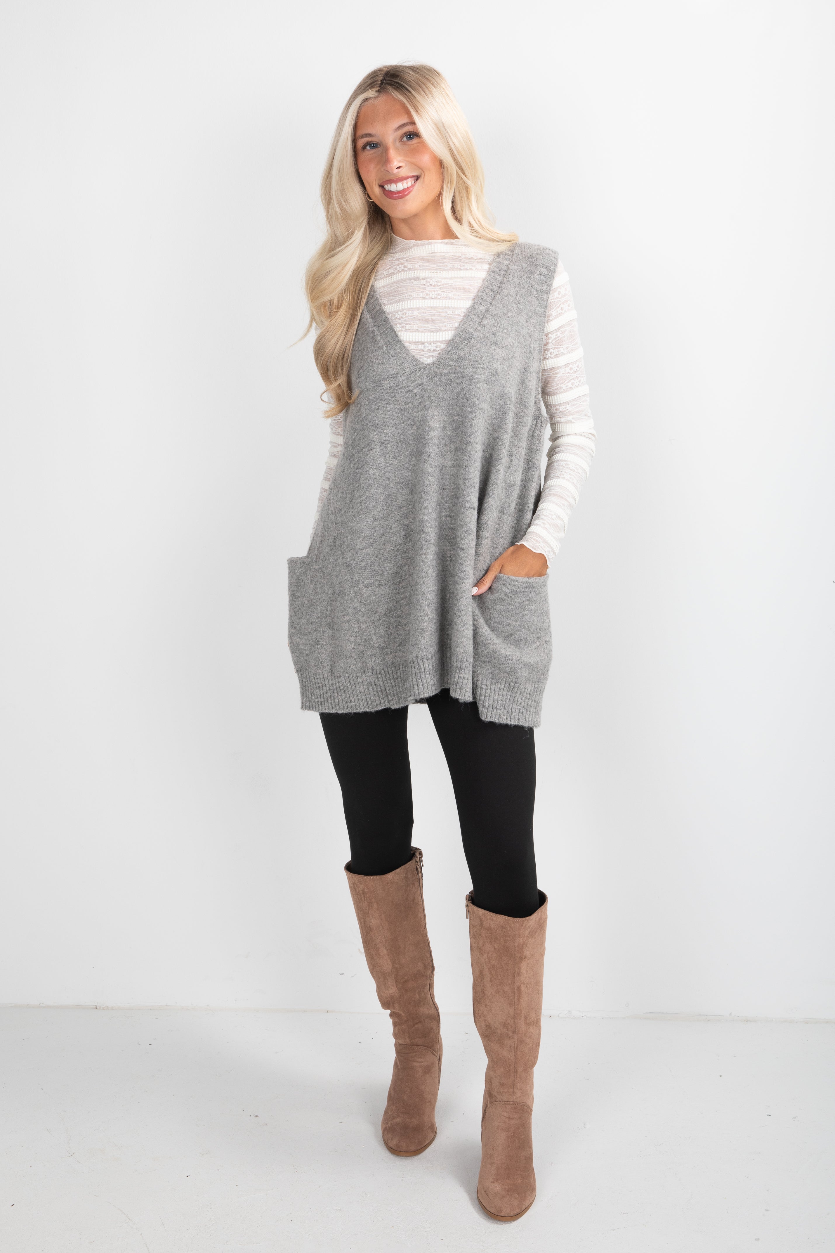 Woman wearing a gray sleeveless sweater, black leggings, and brown knee-high boots on a white background