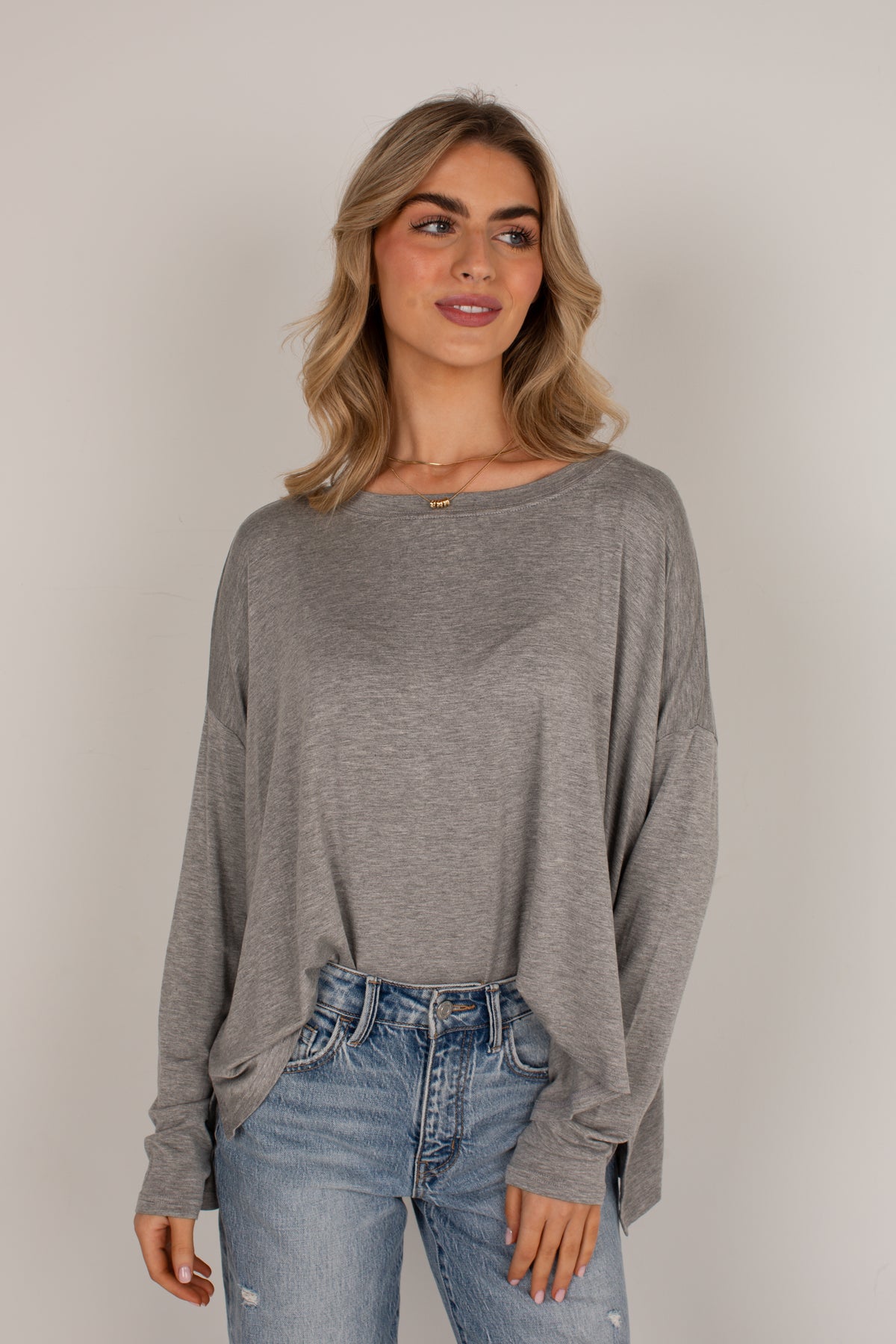 A woman wearing a heather gray long-sleeve top and blue jeans, standing against a plain light gray studio background.