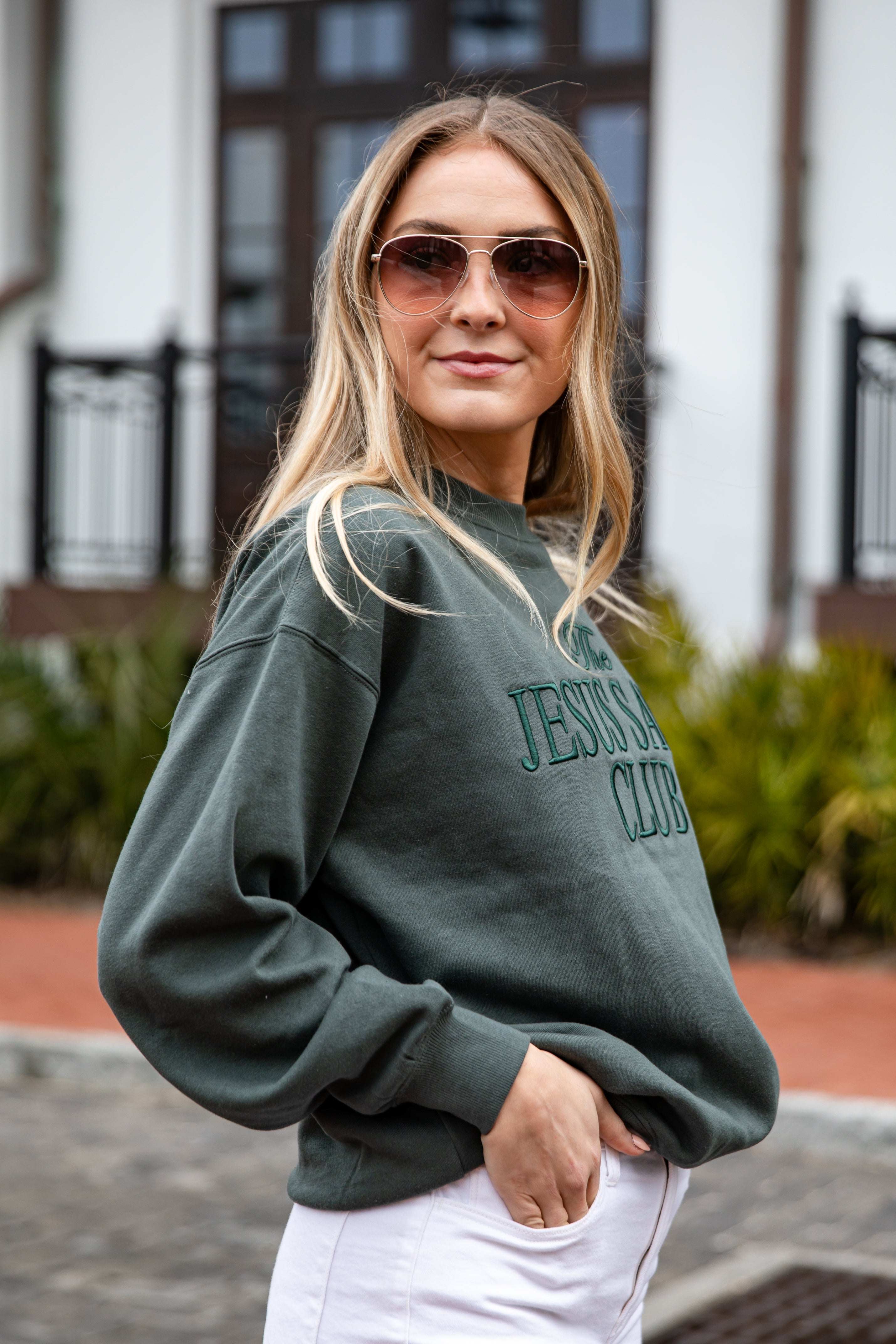 Woman wearing a green sweatshirt with text, sunglasses, and white pants outdoors.