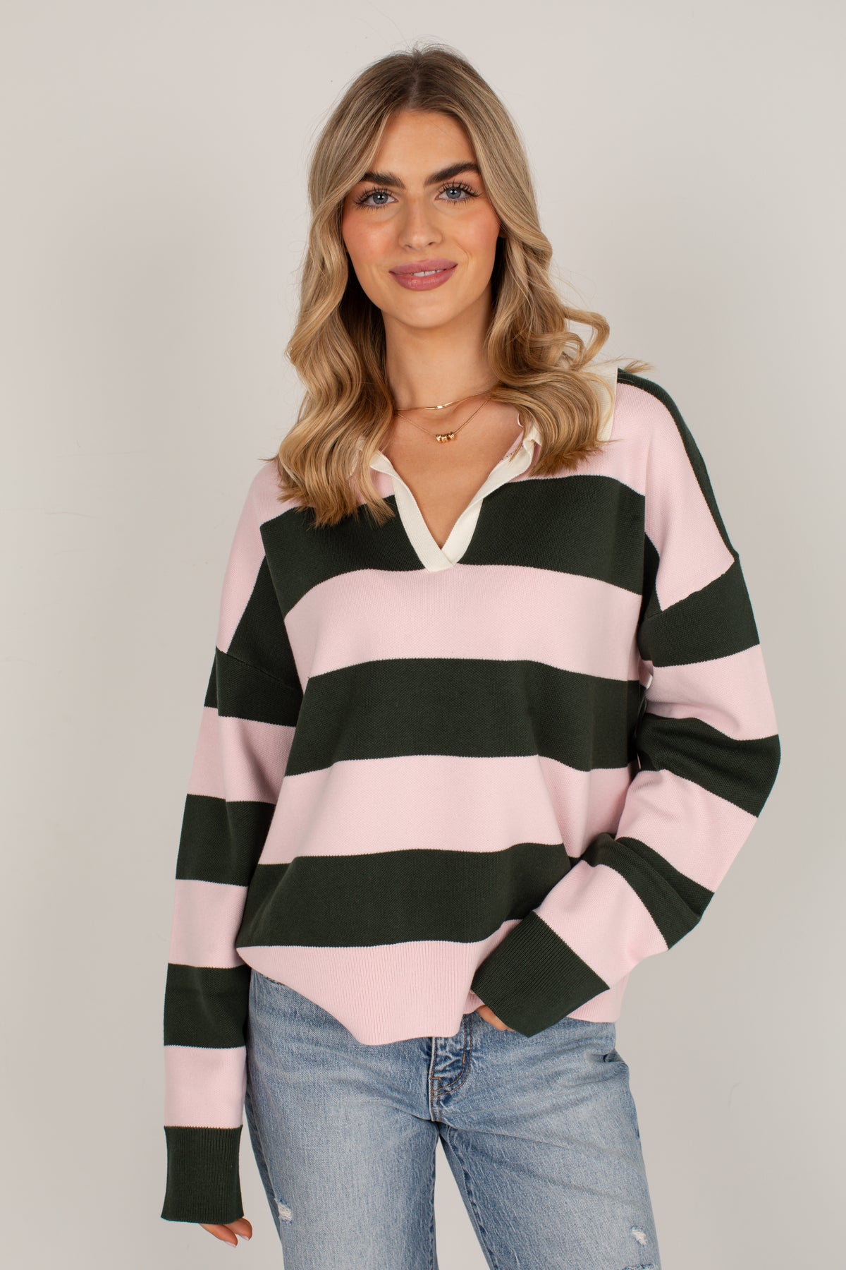 A woman modeling a pink and dark green horizontally striped long-sleeve henley sweater with a white inner placket, paired with light blue jeans, against a solid light gray background.