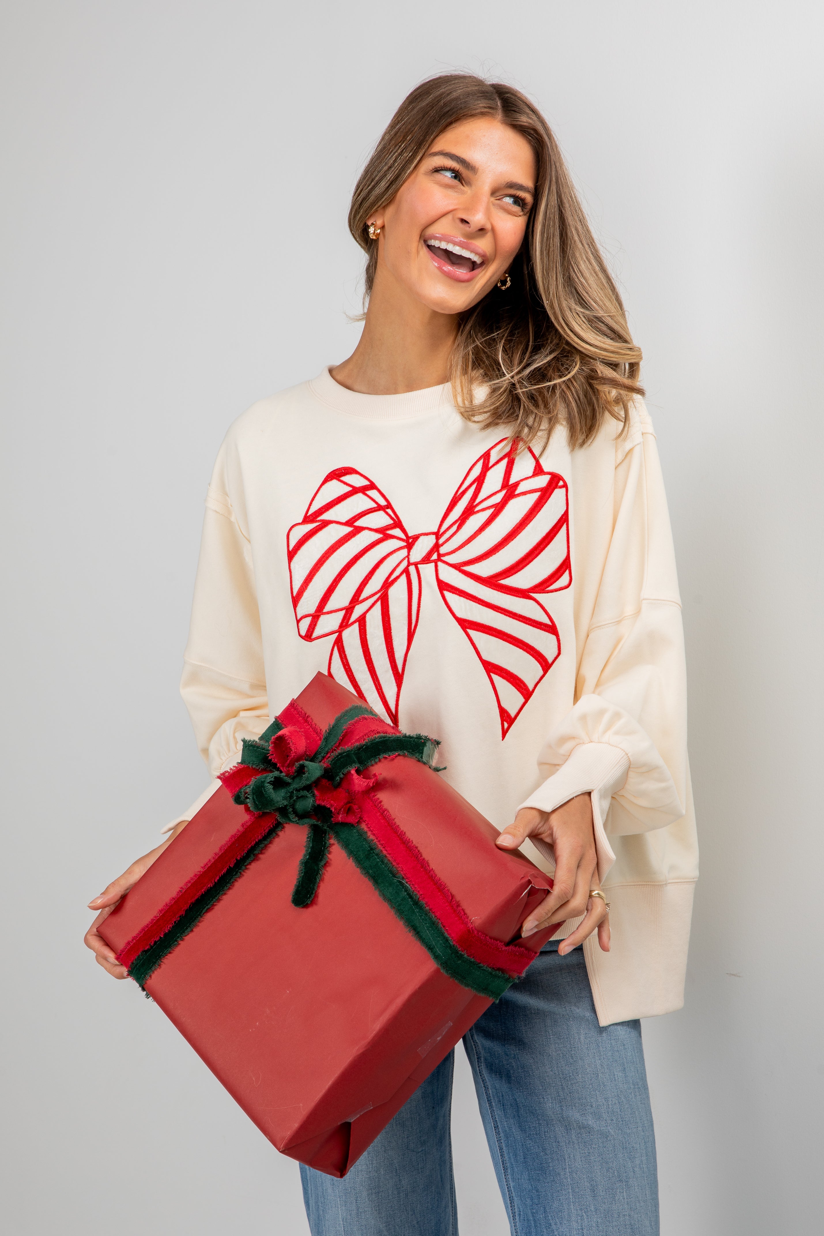 Woman holding a large red gift box with a white sweater featuring a red bow design.