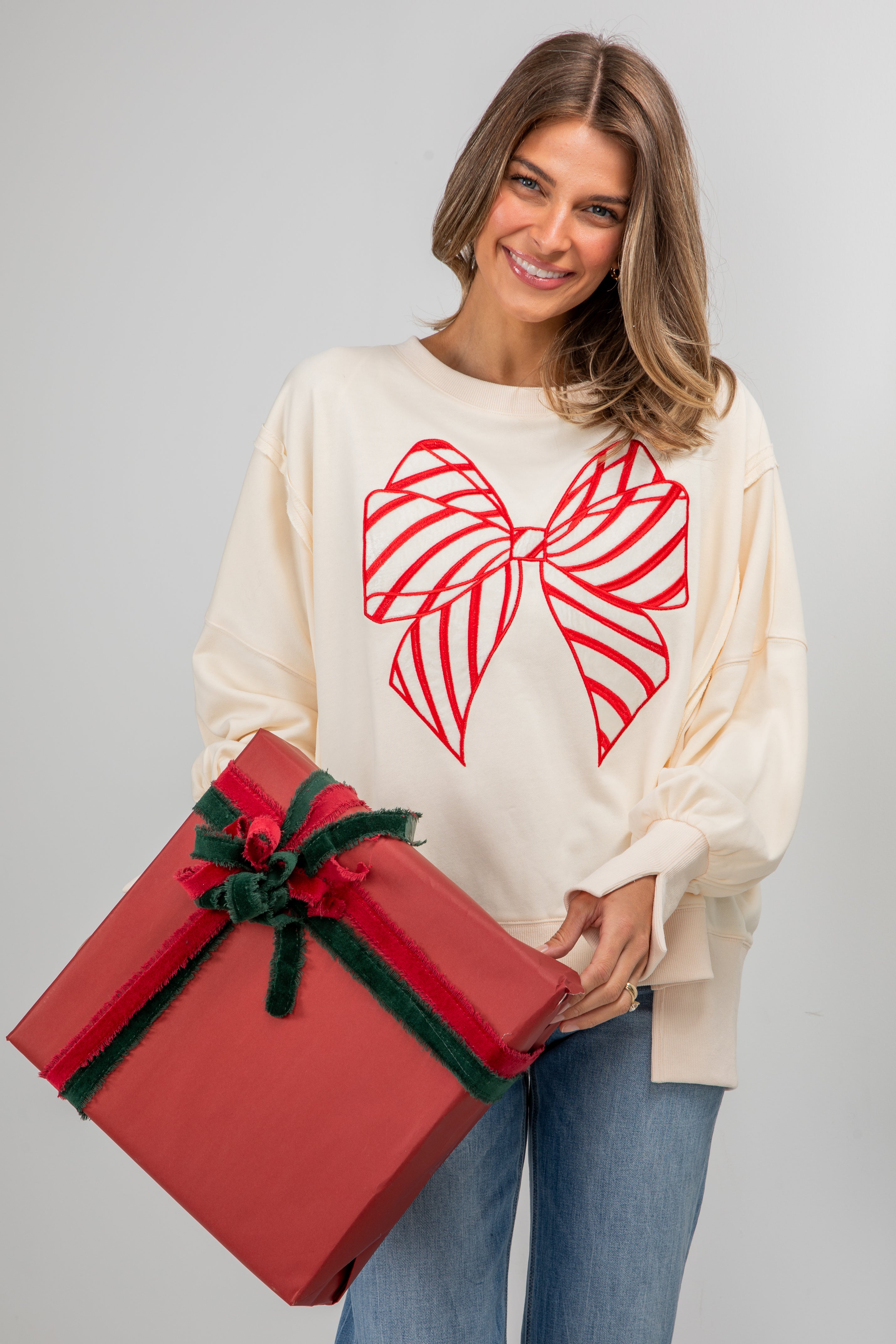 Woman wearing a cream sweater with a red bow design, holding a large red gift box against a gray background