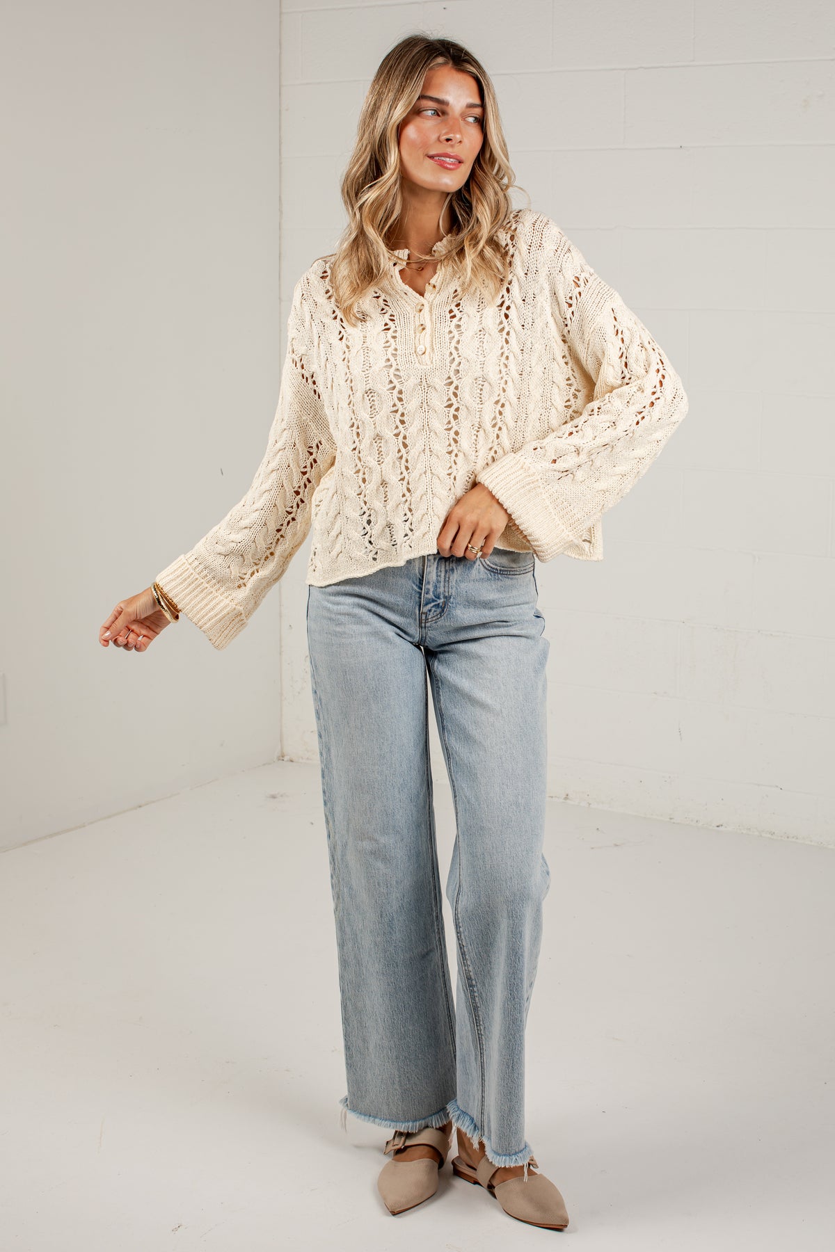 A woman modeling a cream-colored, long-sleeve lace knit blouse paired with light-wash wide-leg jeans and beige pointed flats, standing against a white studio wall.
