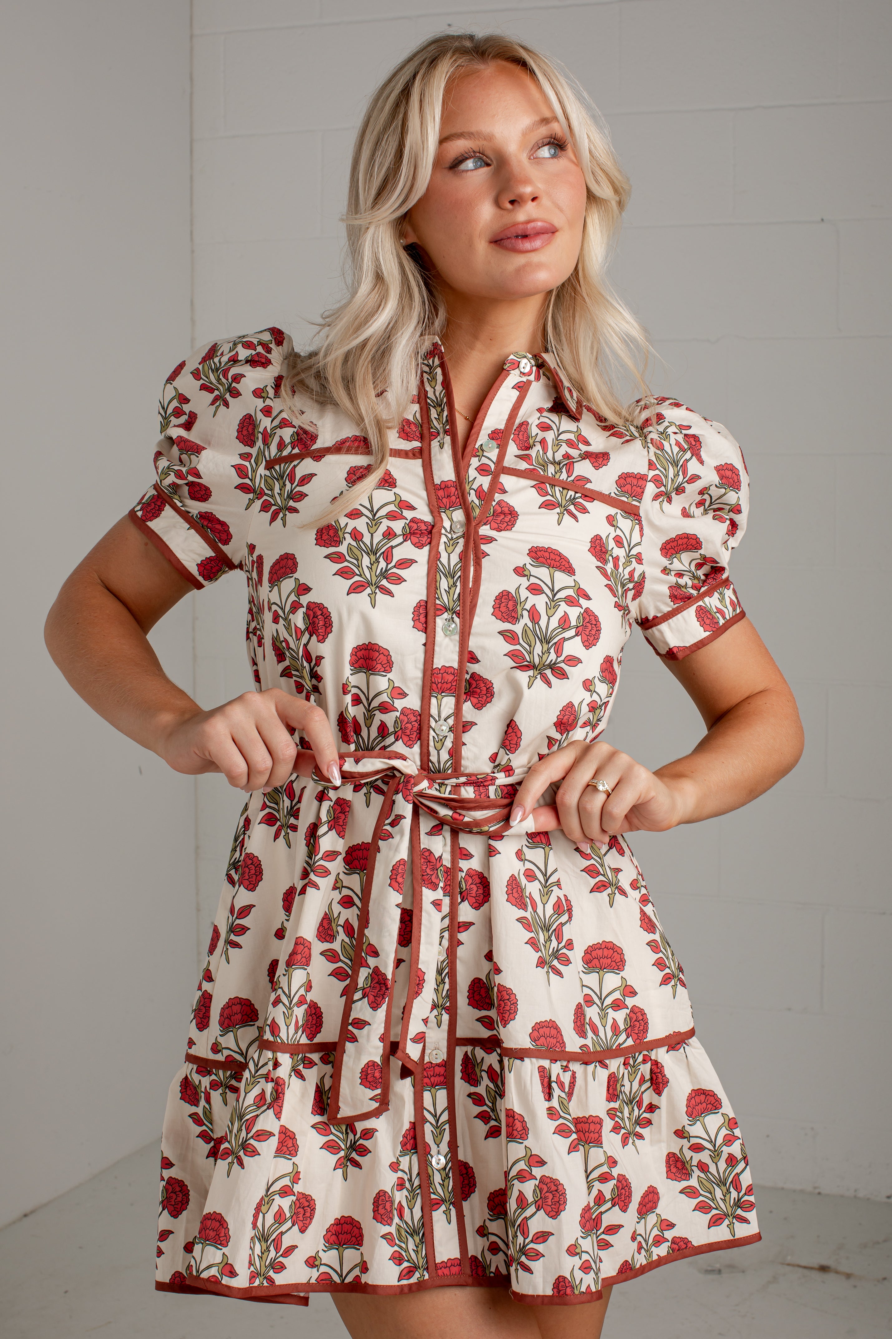 A woman modeling a short-sleeve cream dress with a red and green floral print and a matching tie belt, set against a light gray paneled wall.