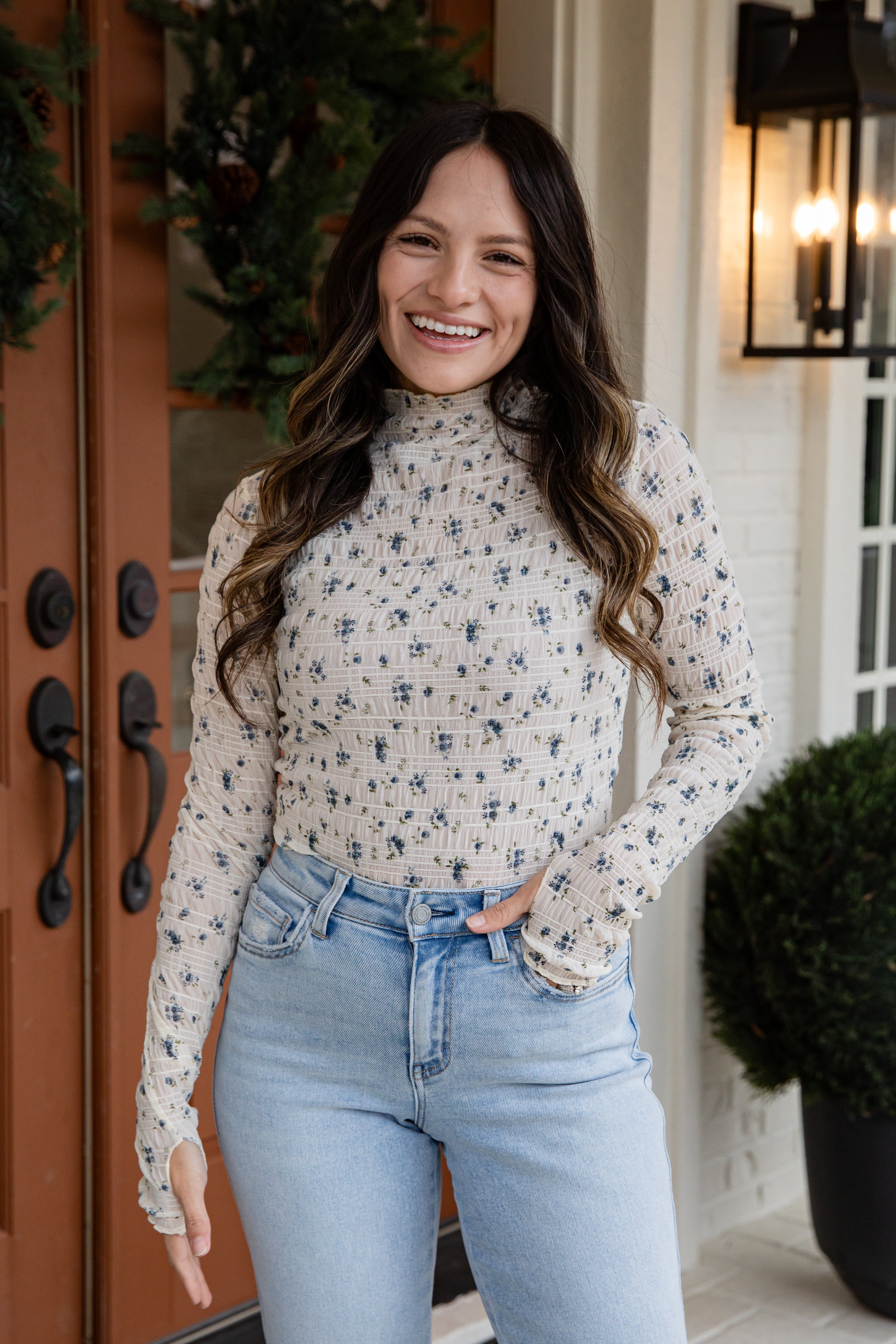 Woman wearing a floral long-sleeve top and jeans standing in front of a door.
