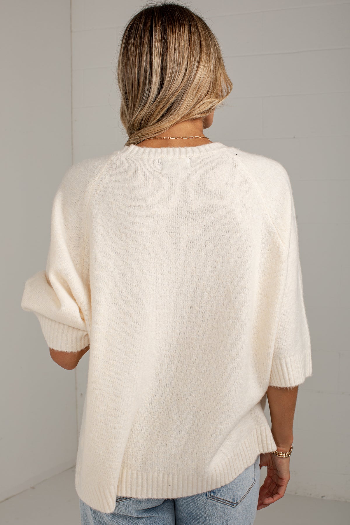 A woman viewed from behind wearing a cream-colored, oversized knit sweater with three-quarter sleeves and light blue jeans, set against a white paneled wall.
