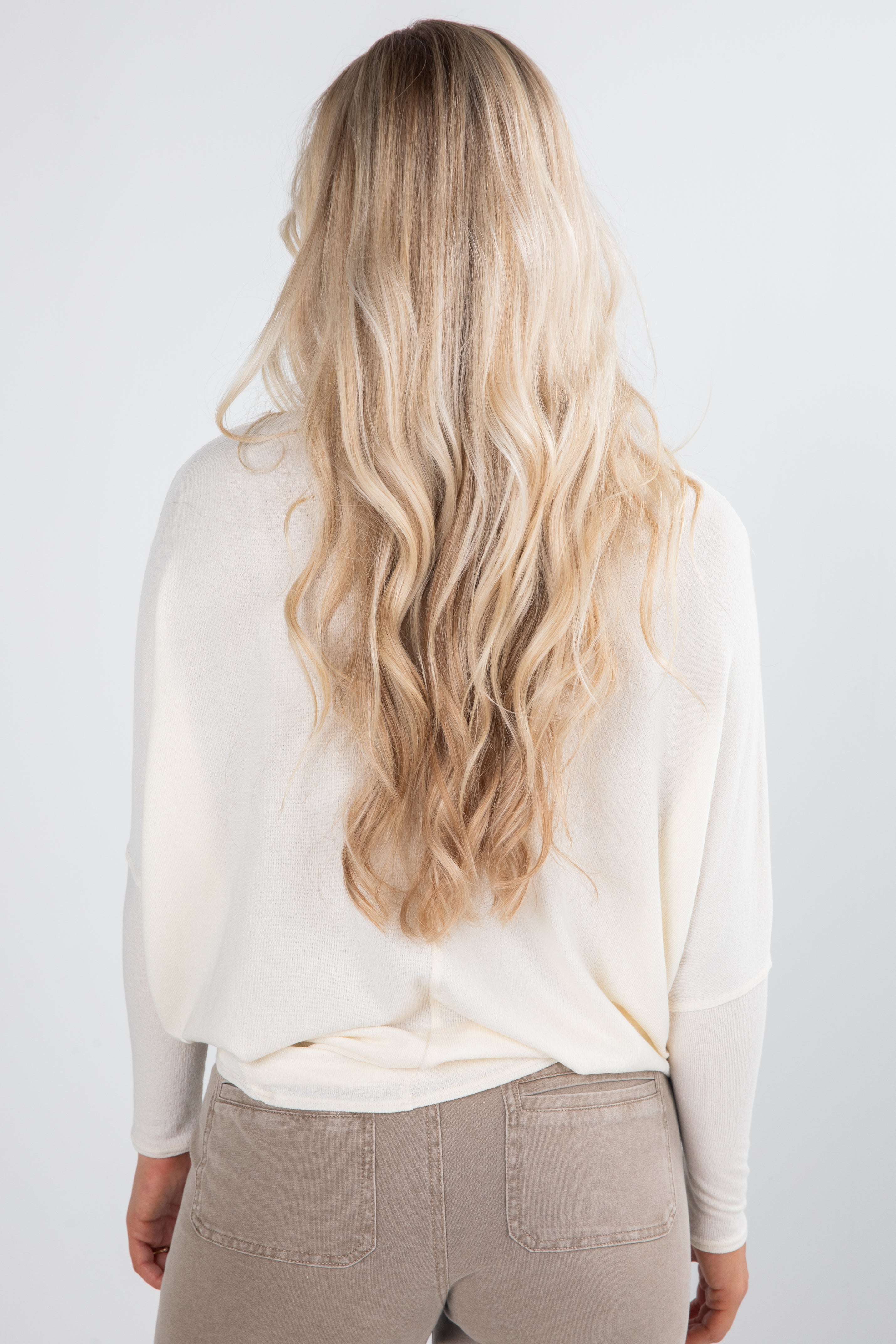 Person with long blonde hair wearing a white shirt and beige pants on a plain background