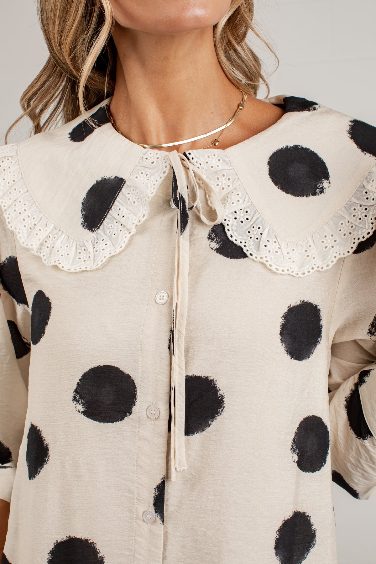 A close-up of a woman wearing a cream-colored blouse with large black polka dots and a large white eyelet Peter Pan collar with a tie detail.