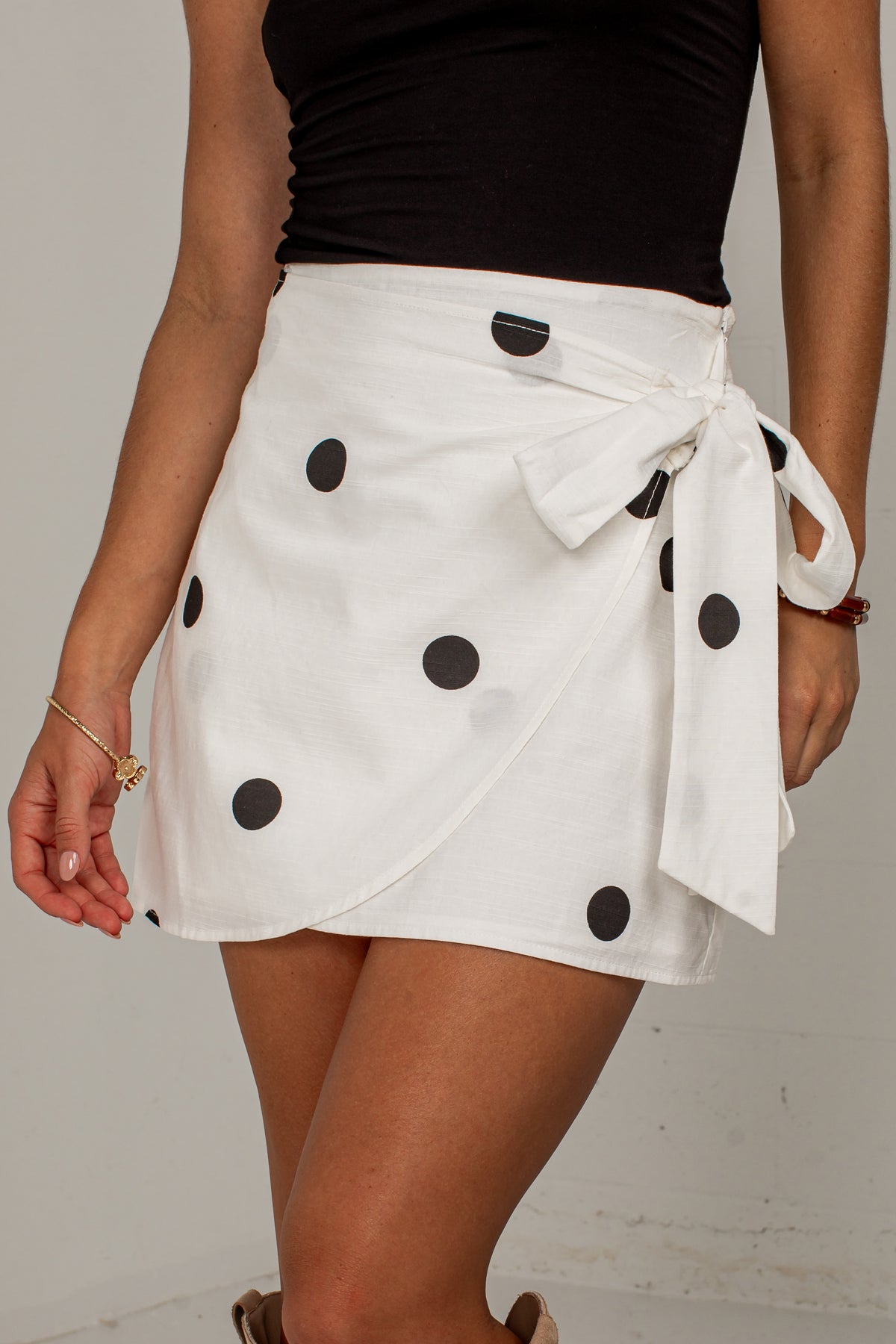 A woman wearing a white wrap-style mini skirt with black polka dots and a tied waist, paired with a black top against a plain light background.