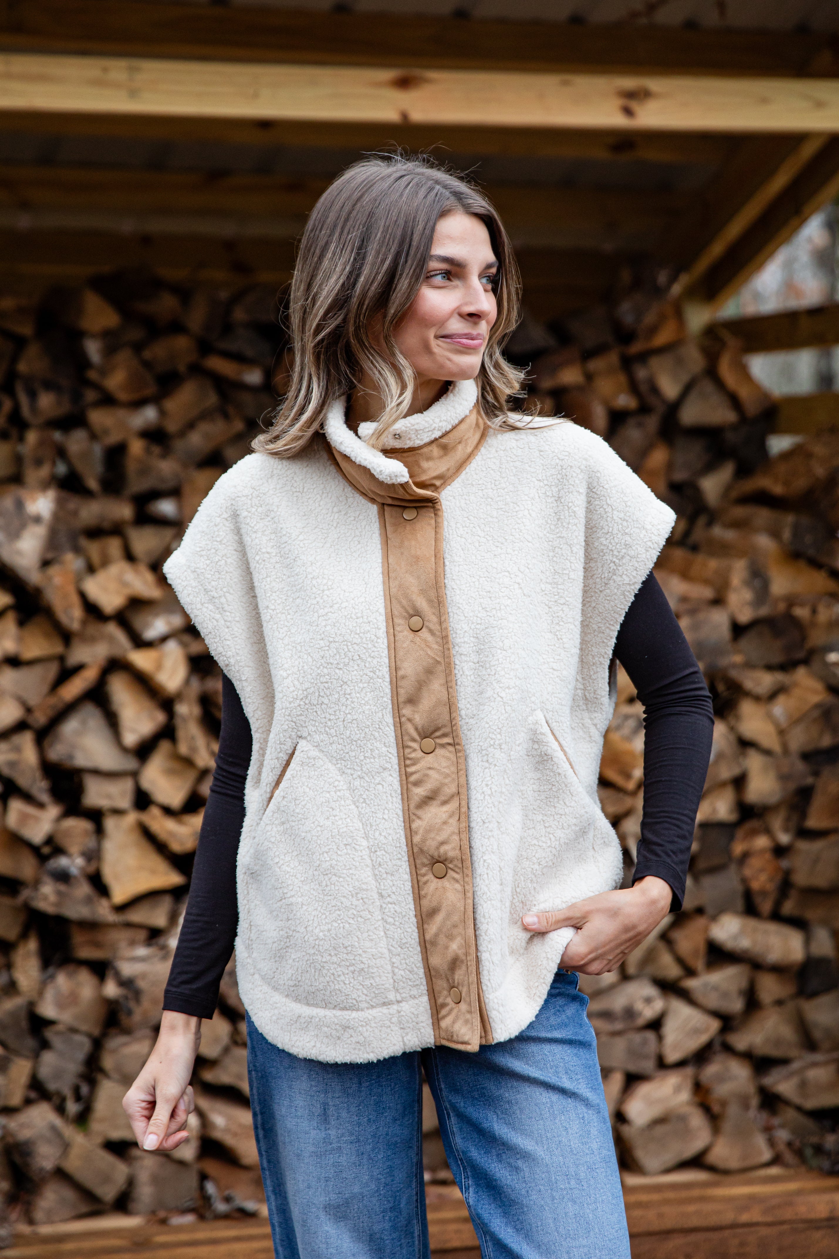 Woman wearing a cream-colored vest with a brown front panel in front of stacked firewood.