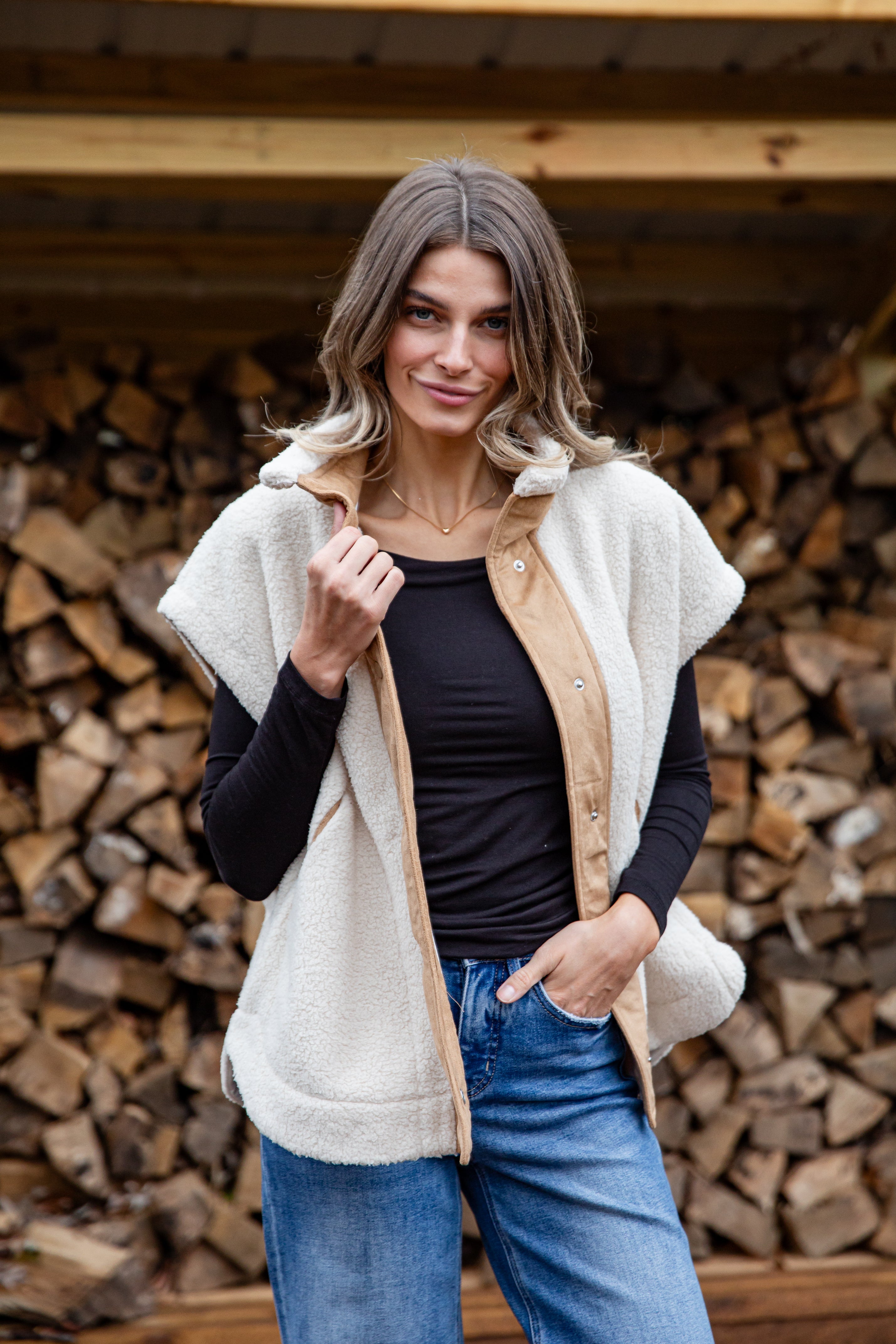 Woman wearing a beige vest over a black top and blue jeans, standing in front of stacked firewood.