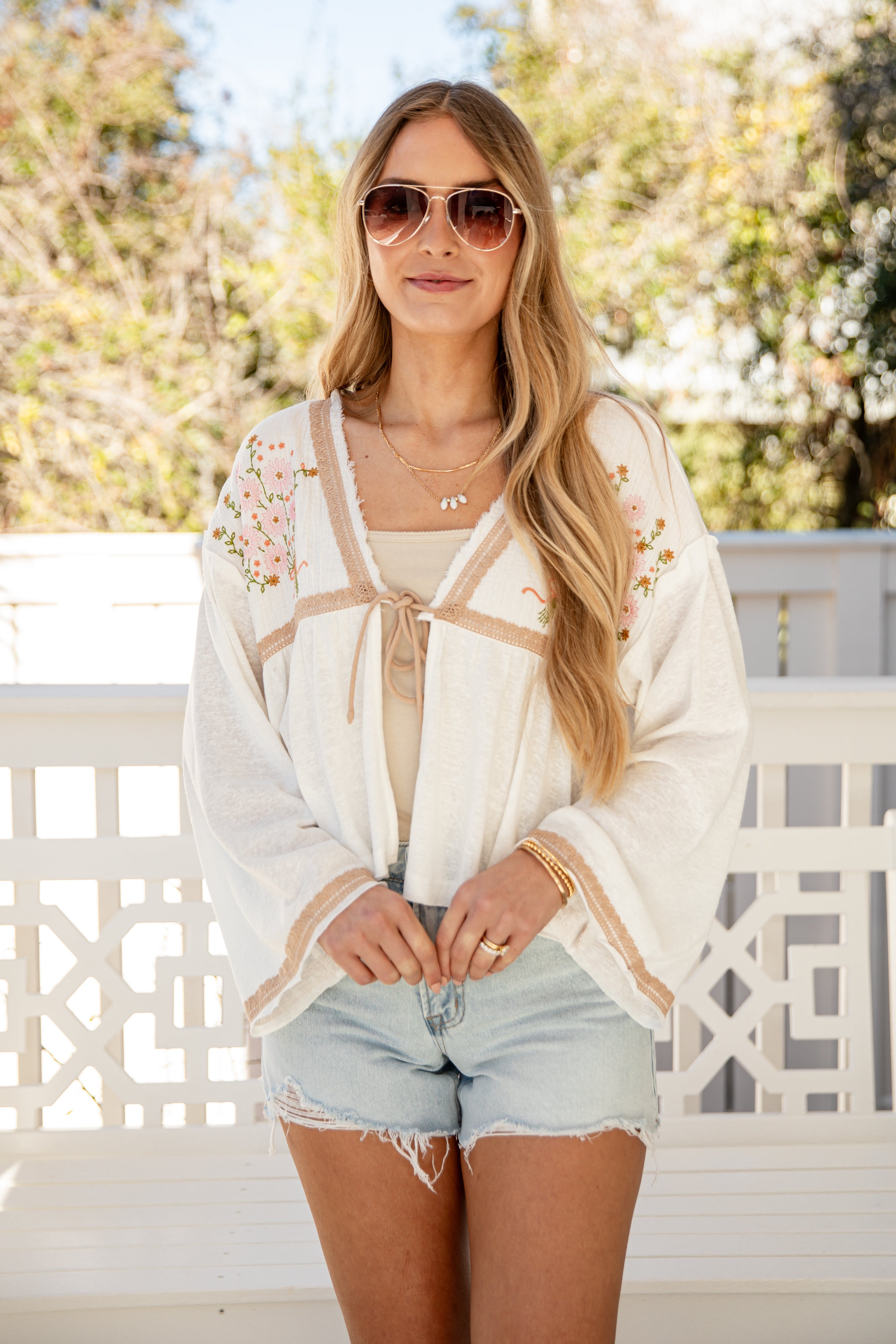 Woman wearing a white cardigan with floral details, sunglasses, and denim shorts on a balcony.