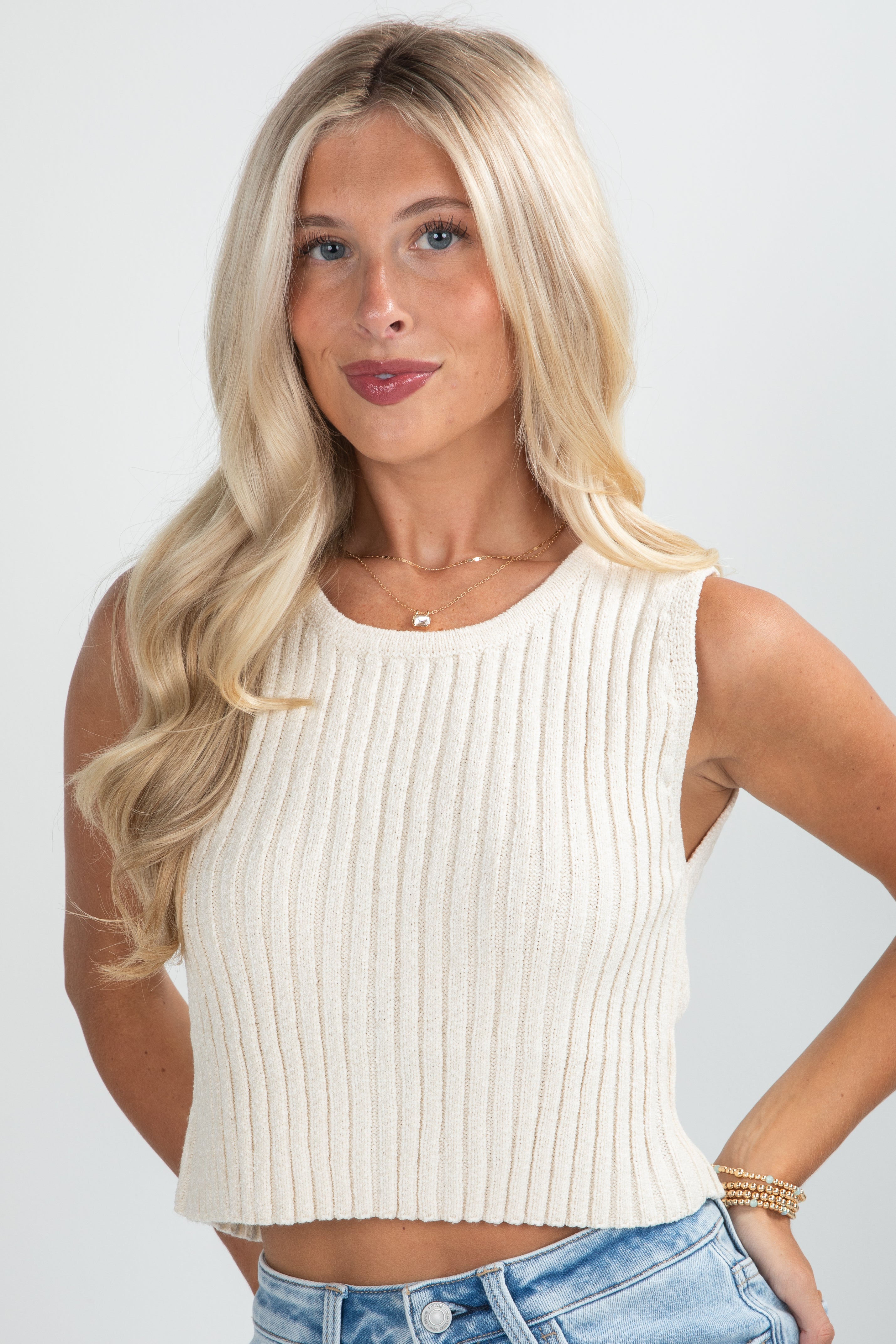 Woman wearing a white ribbed tank top and blue jeans against a plain background