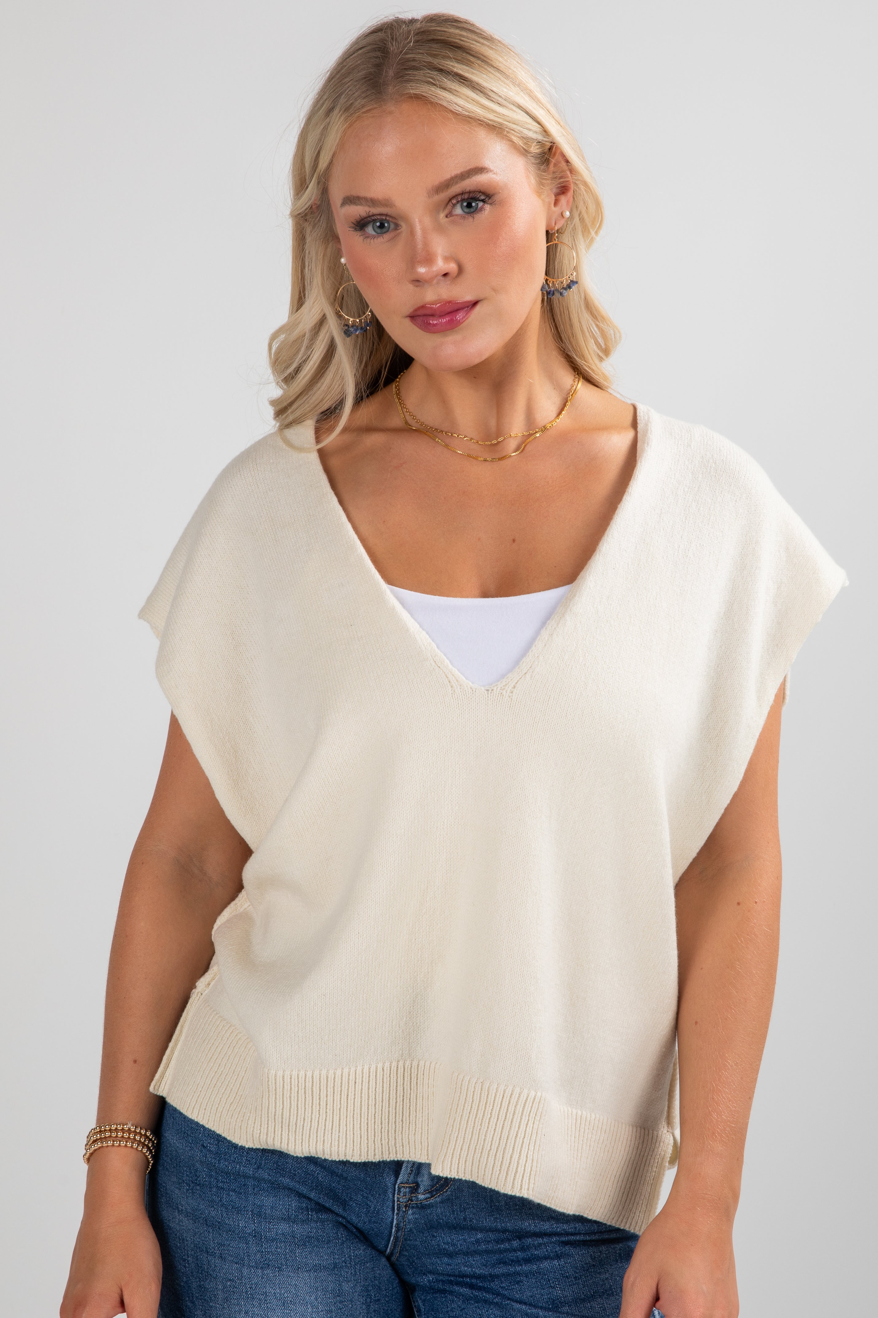 Woman wearing a beige sweater over a white shirt with blue jeans on a plain background