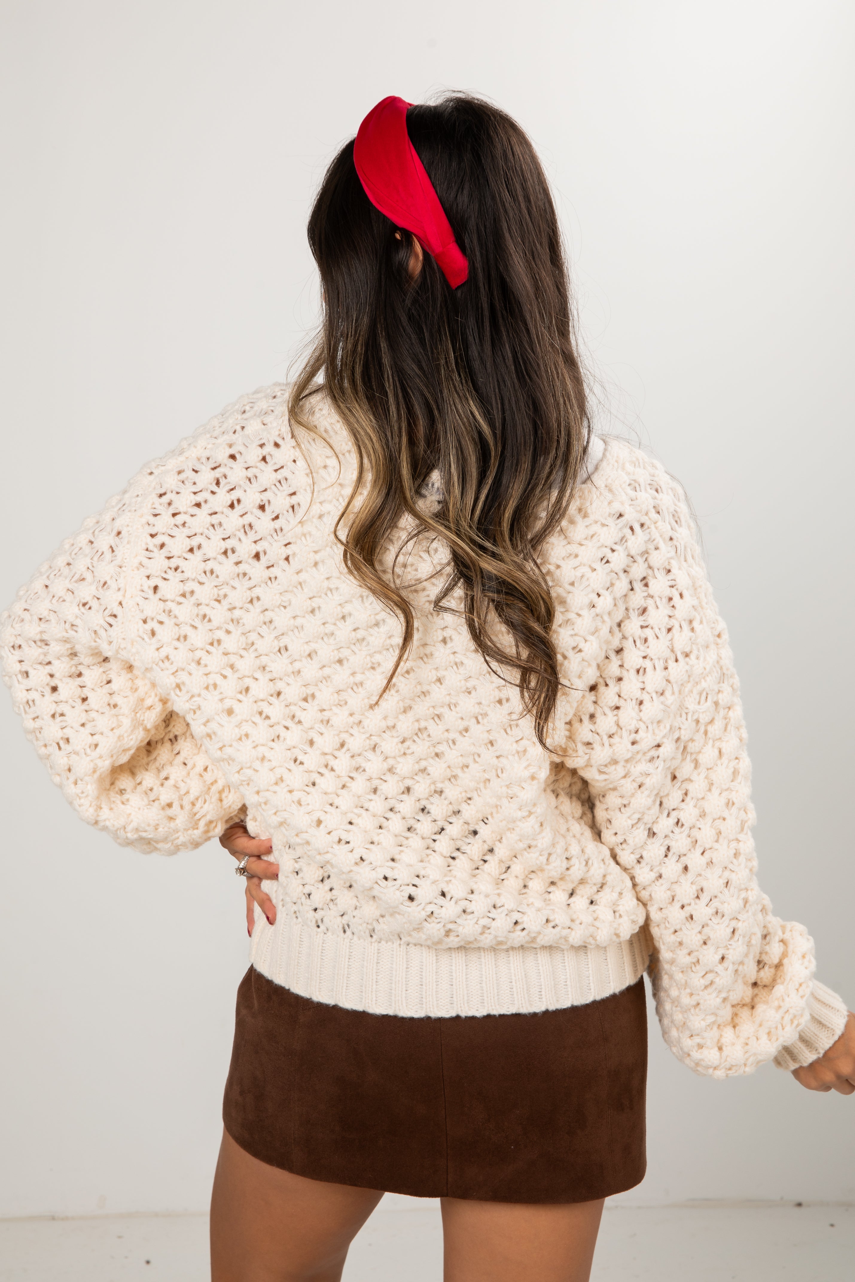 Woman wearing a beige chunky knit sweater and brown skirt with a red headband against a white background