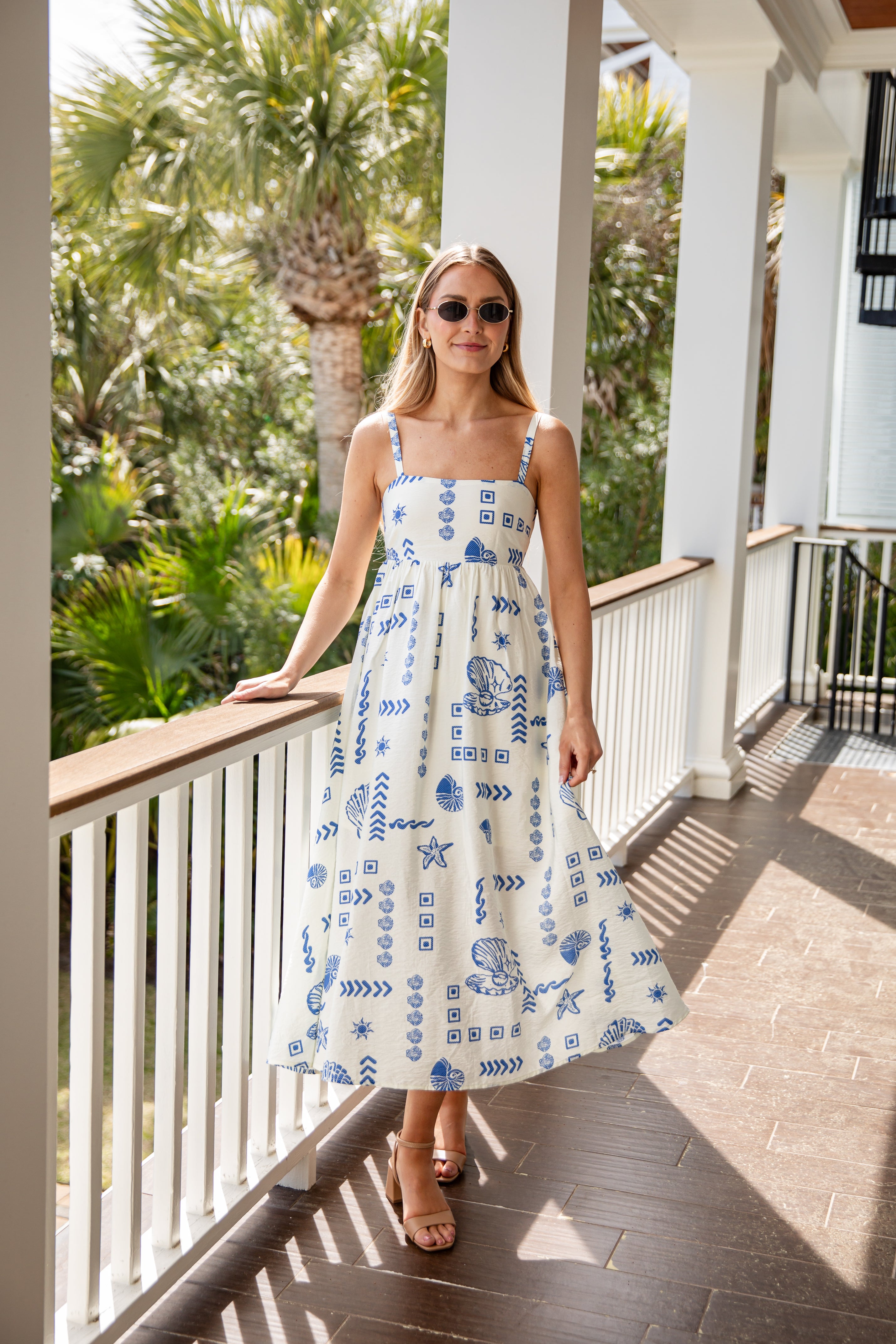Woman in a white dress with blue patterns standing on a porch with palm trees in the background