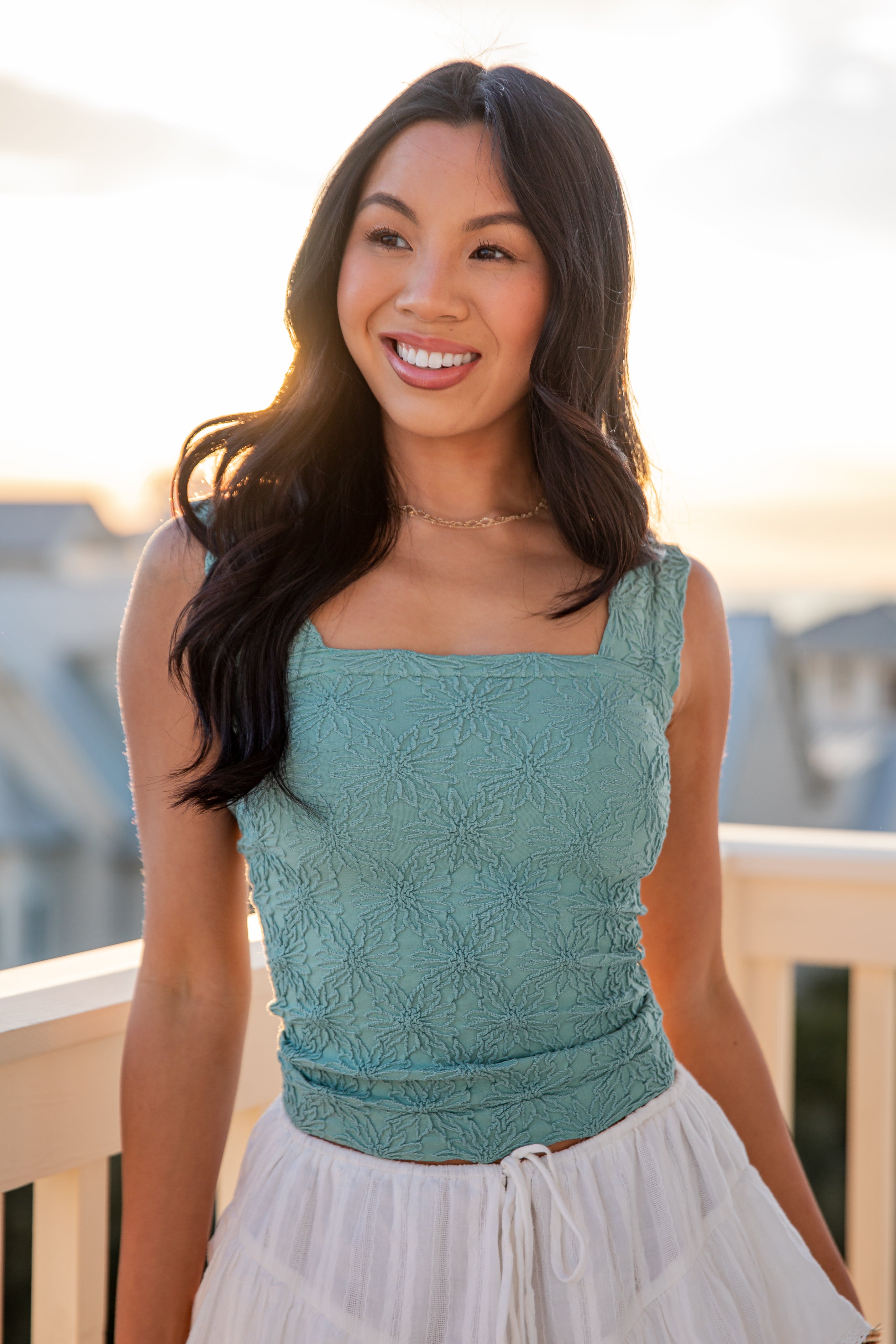 Woman wearing a teal lace top and white skirt on a balcony with a blurred background