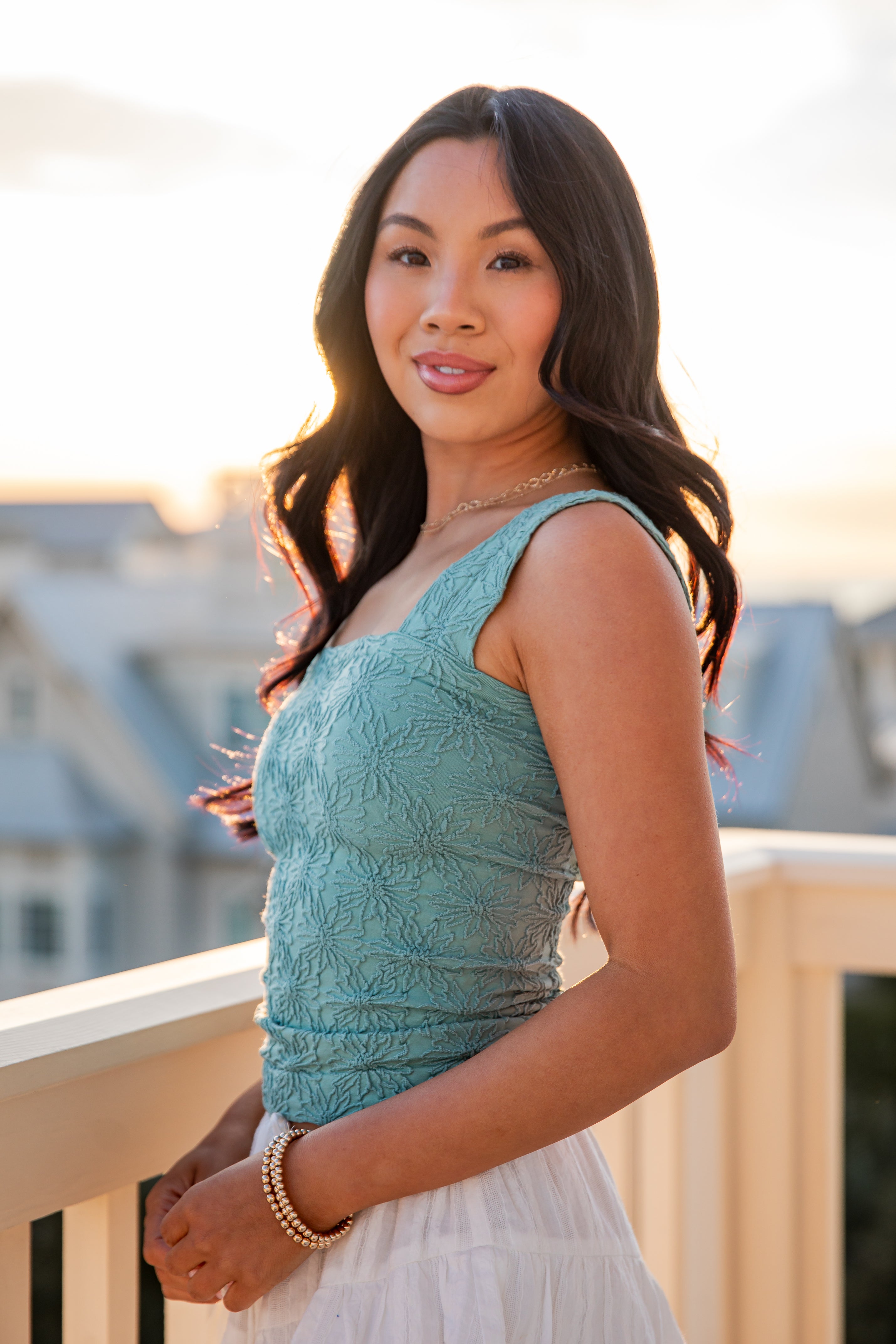 Woman in a teal top and white skirt standing on a balcony with a blurred cityscape background