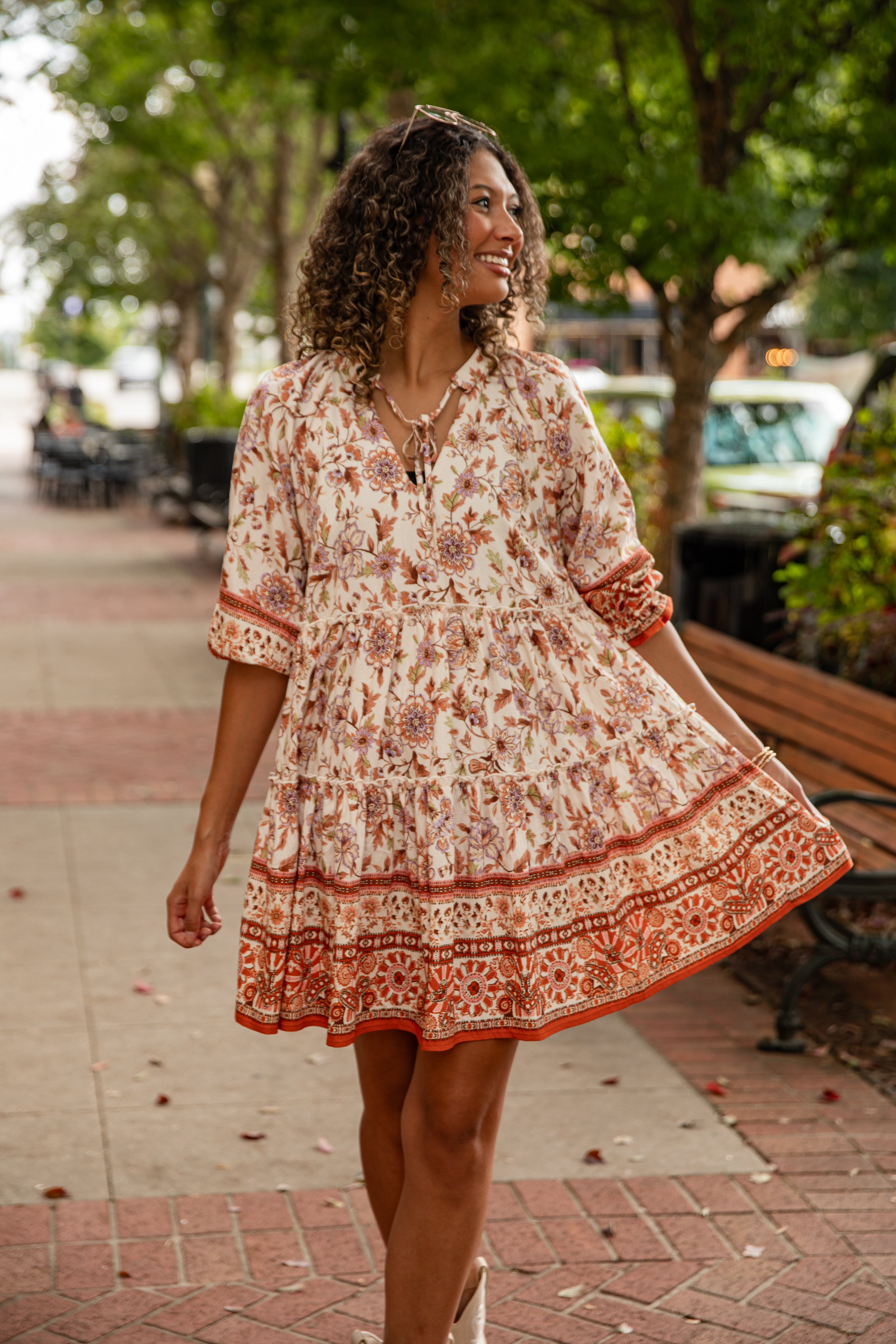 Woman in a patterned dress standing on a sidewalk with trees and benches in the background